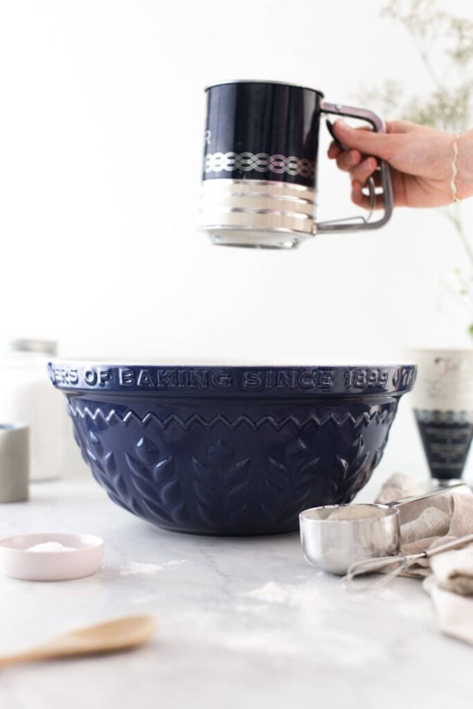 A hand using a flour sifter to sift flour into a bowl.