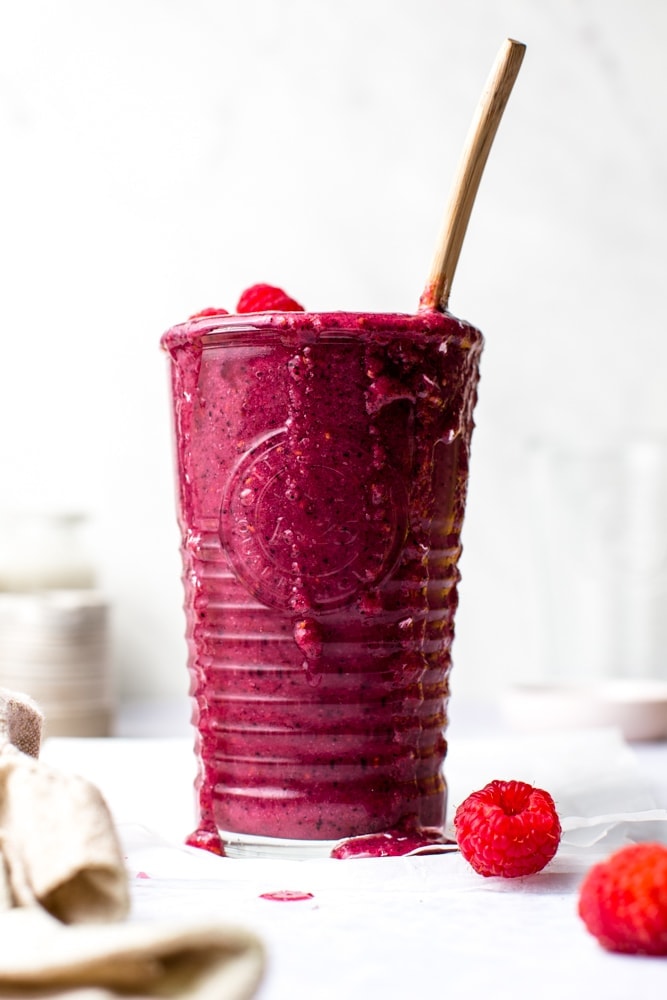 A glass of smoothie with raspberries scattered around. 