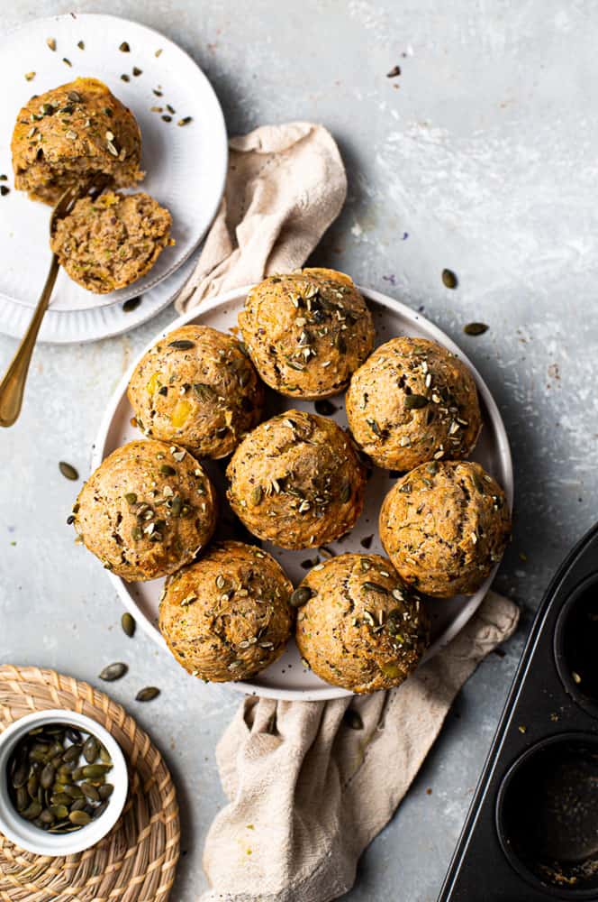 Eight savoury vegan muffins placed on a large round plate with a napkin, pumpkin seeds and more small plates placed around them. 