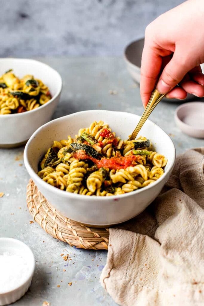 A hand stirring a bowl of vegan pesto pasta salad. 