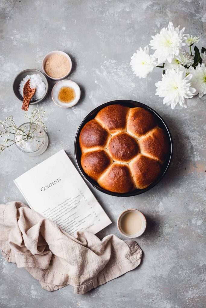 A flatlay scene containing a round loaf of dinner rolls, book, flowers, napking and ingredients. 