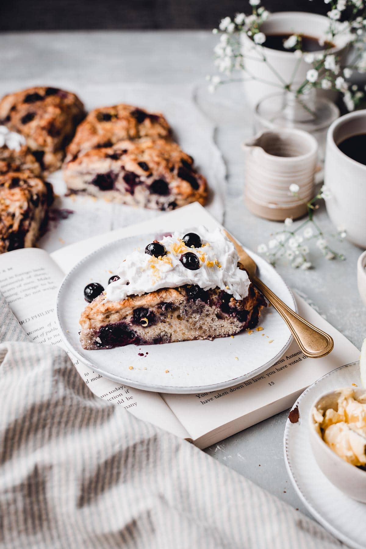 A side view of a scone on a plate placed on top of an open book with flowers, props and more scones in the background. 