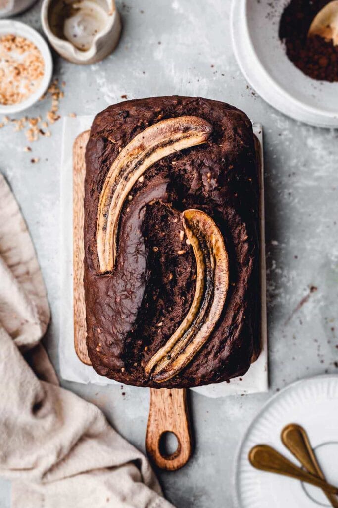 A large banana bread on a chopping board surrounded by plates and toppings. 