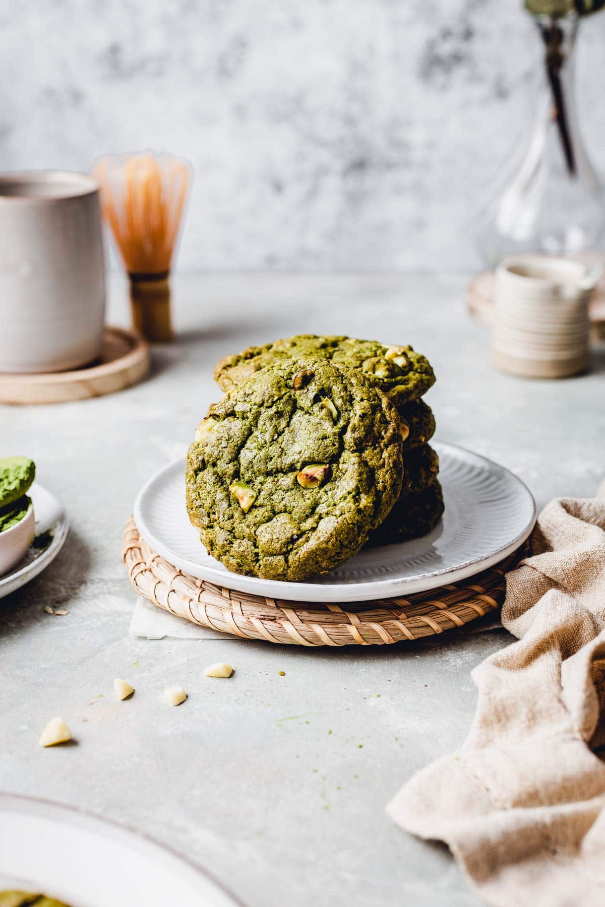 A stack of vegan matcha cookies on a small round plate surrounded by napkins and chocolate chunks. 