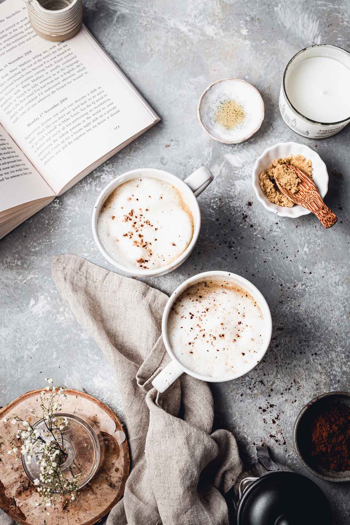 An overhead view of a table with two latte mugs, book and various props laid around it. 