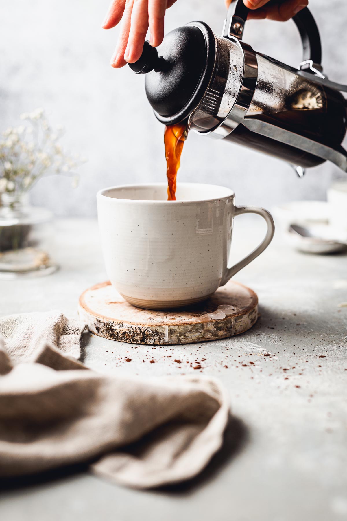 A hand pouring coffee from a French press into a mug placed on a wooden coaster.