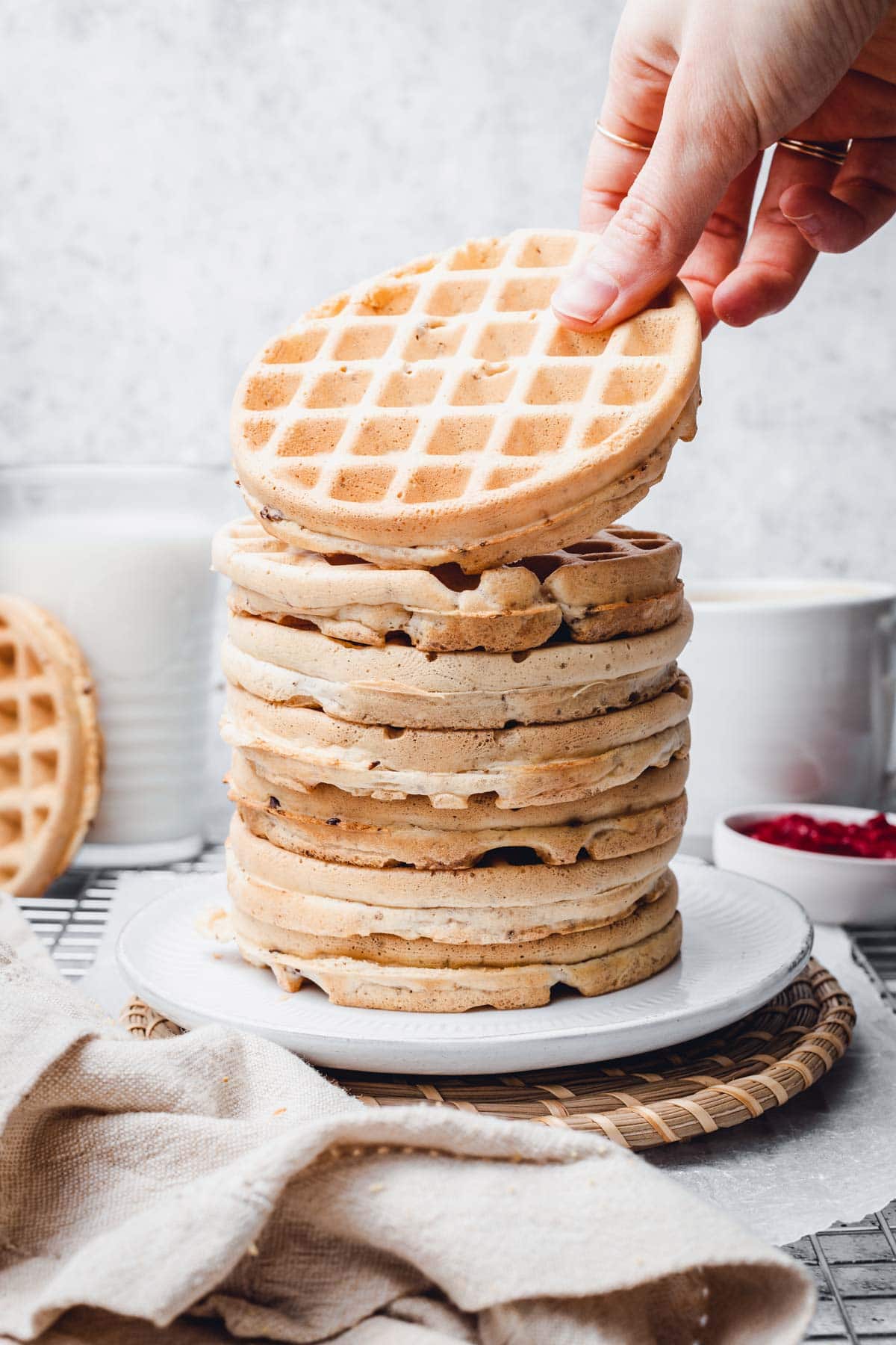 A hand reaching out for a waffle on top of a large waffle stack placed on a round plate.