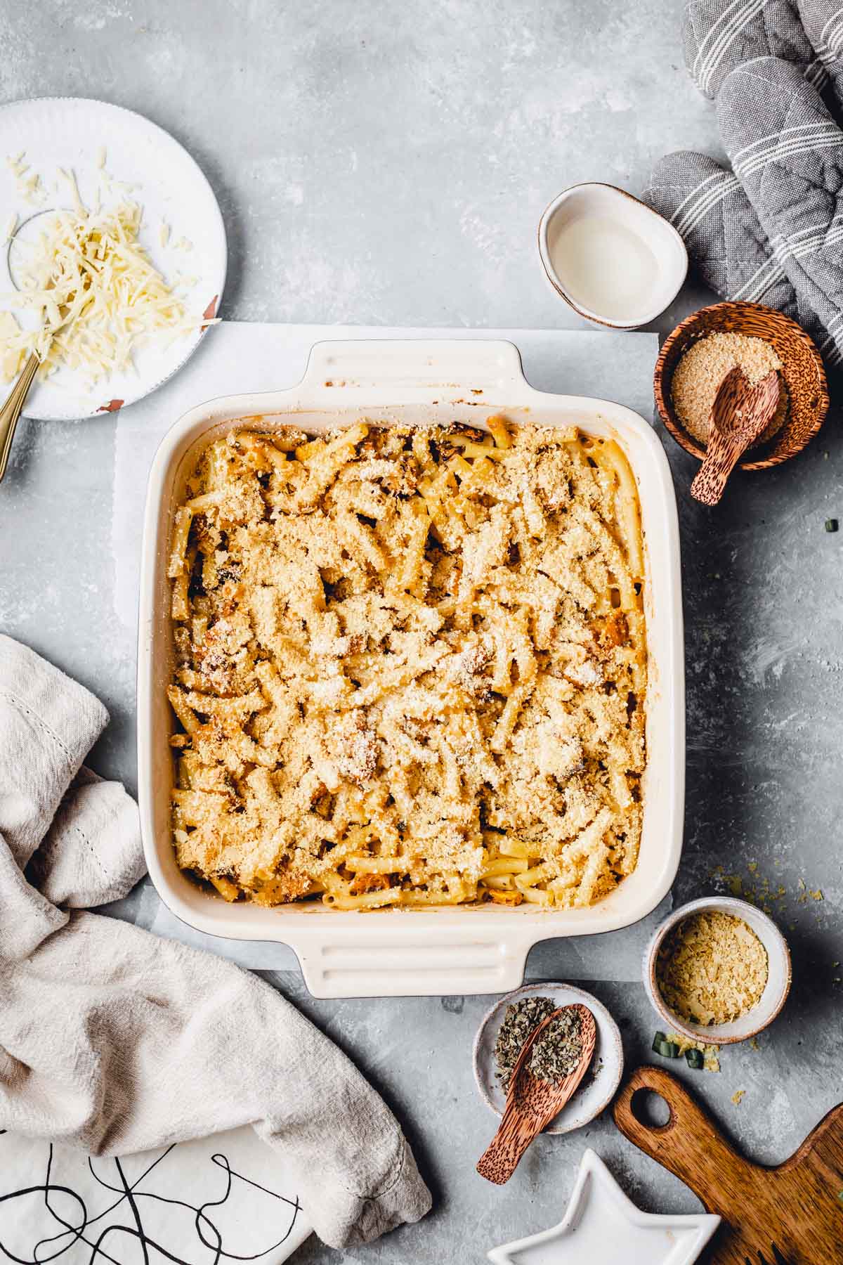 Baked mac and cheese in a rectangular tray surrounded by various small bowls, a linen and a wooden board. 