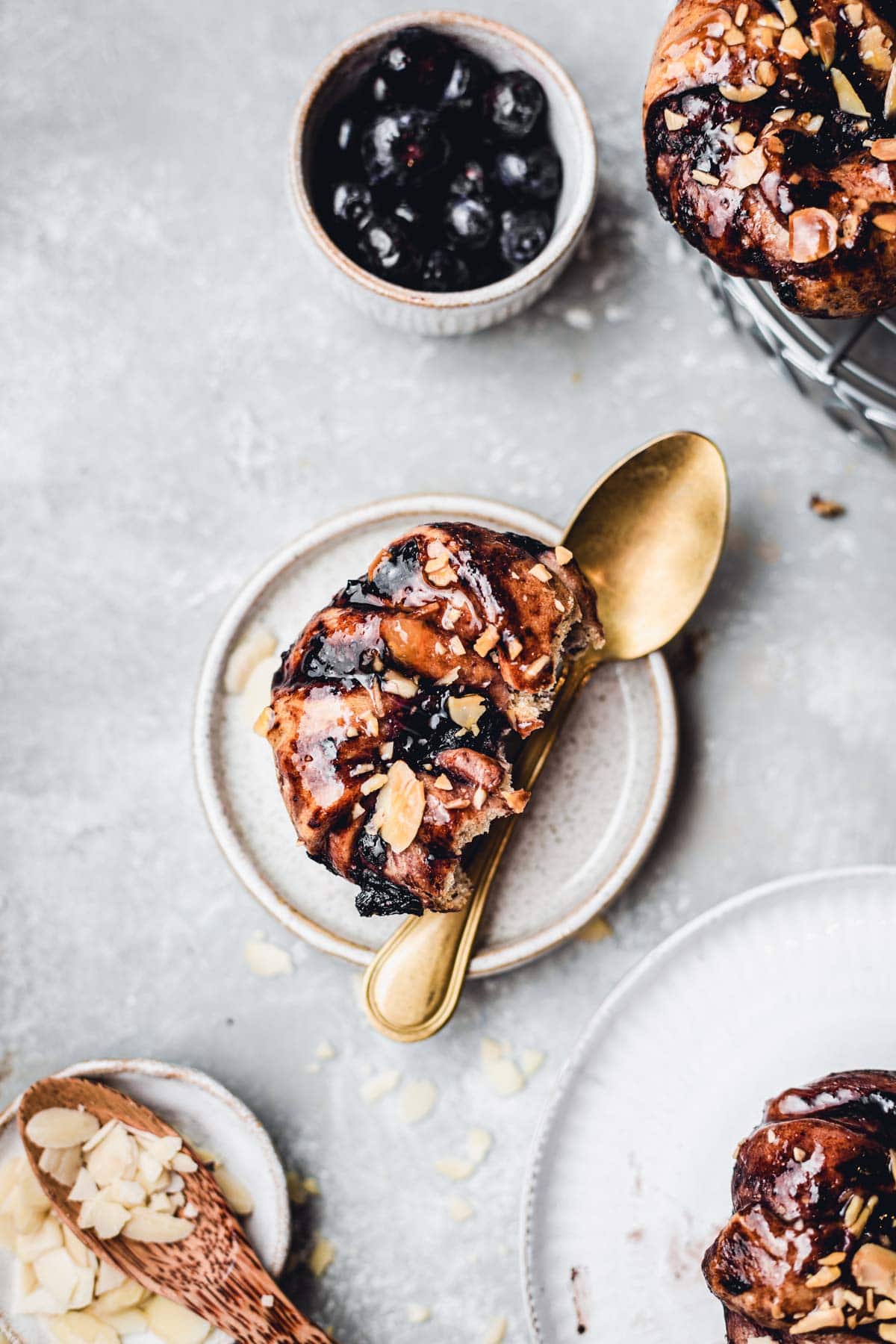 An overhead view of one blueberry cinnamon roll on a small plate with a golden spoon next to it. 