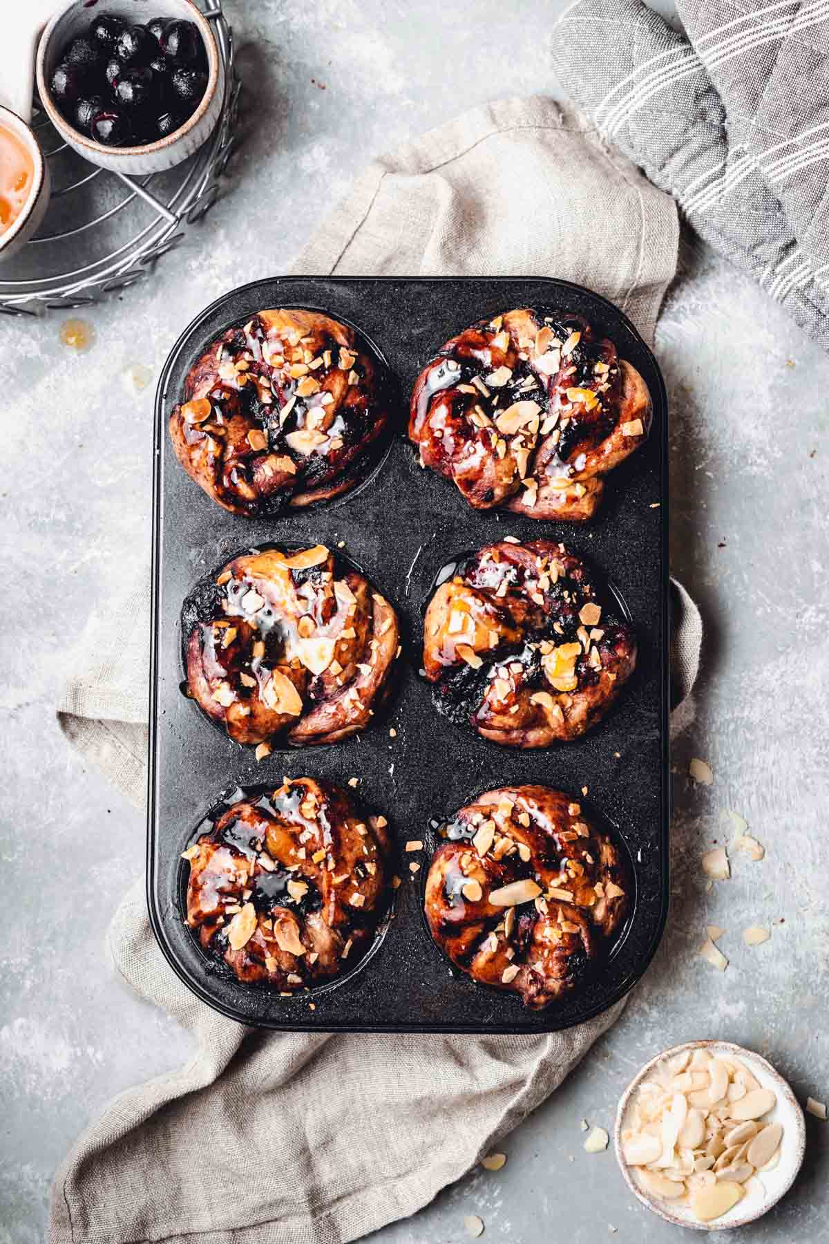 A muffin tray filled with blueberry rolls with a piece of linen and almonds scattered around. 