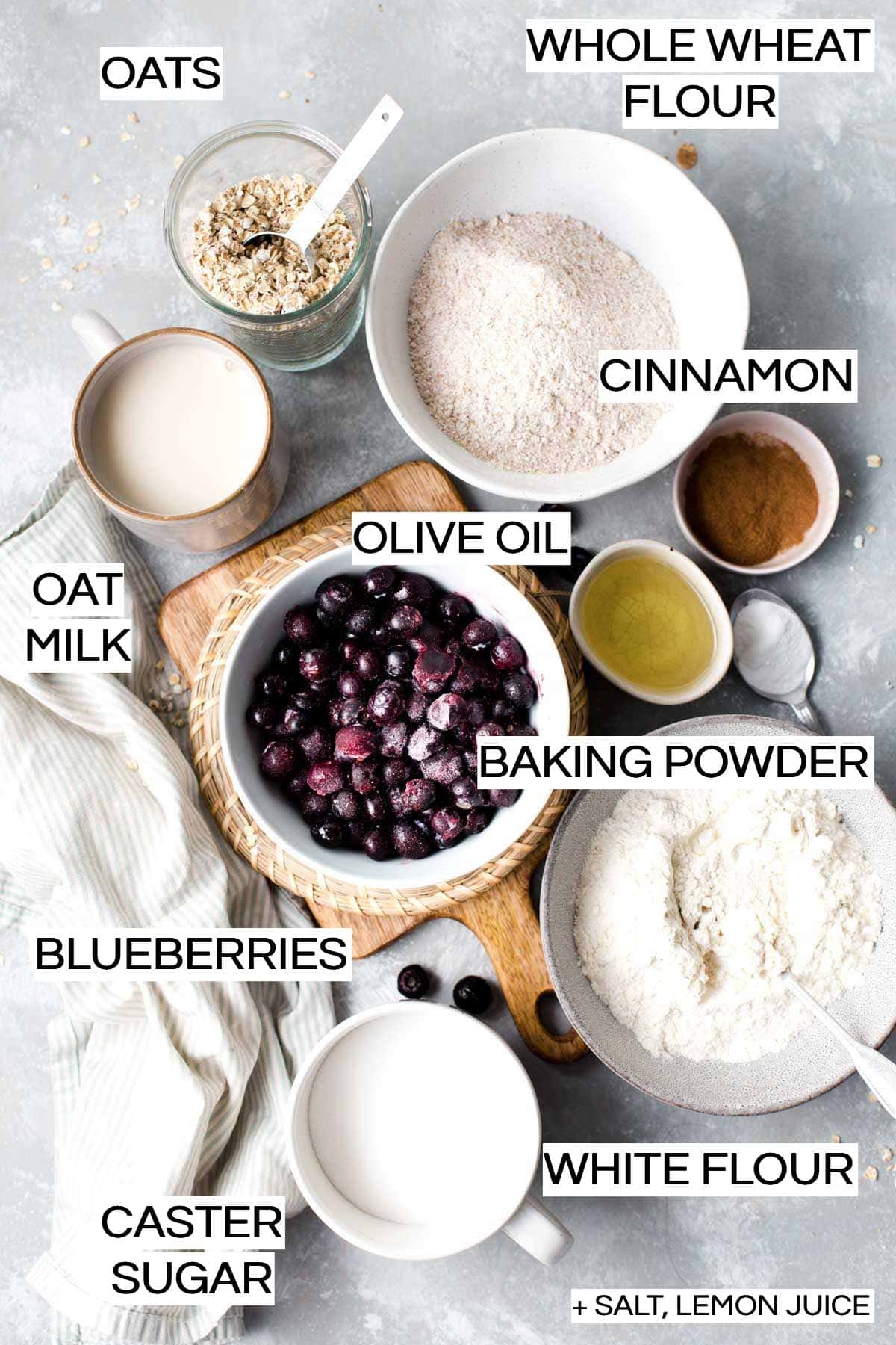 Flour, oats, blueberries, oil and cinnamon laid out in cups and bowls on a flat surface. 