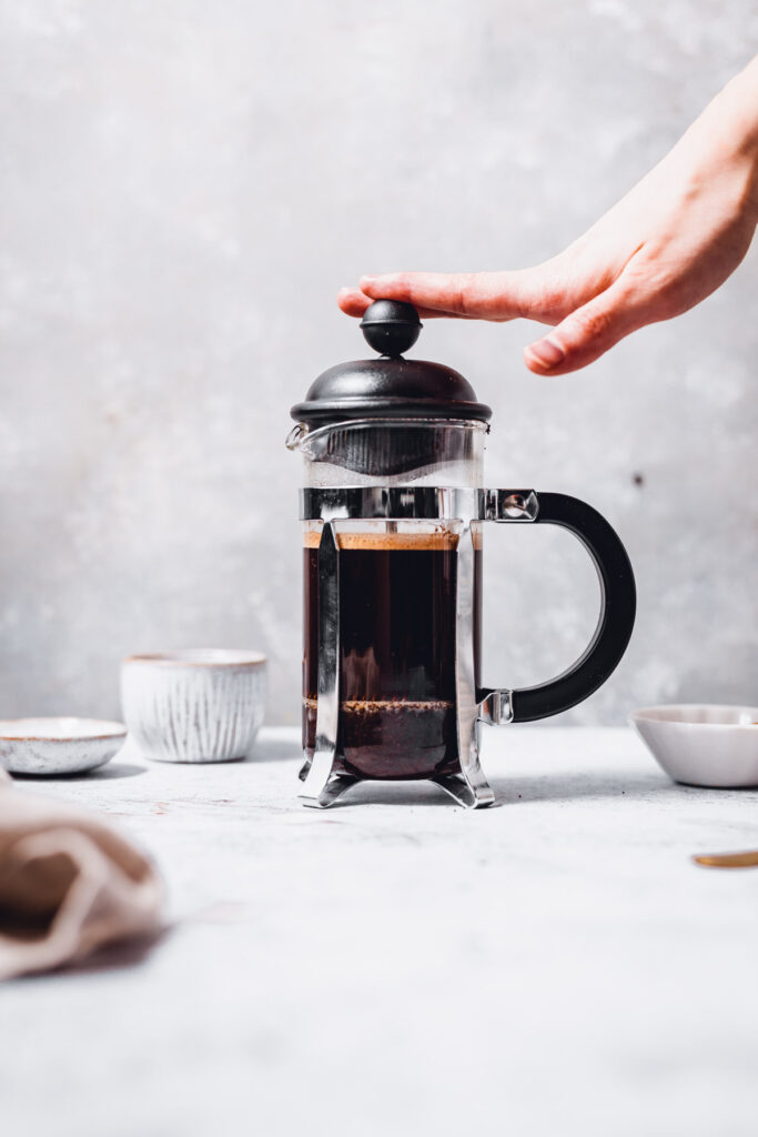 A hand pressing down on a French press filled with coffee. 