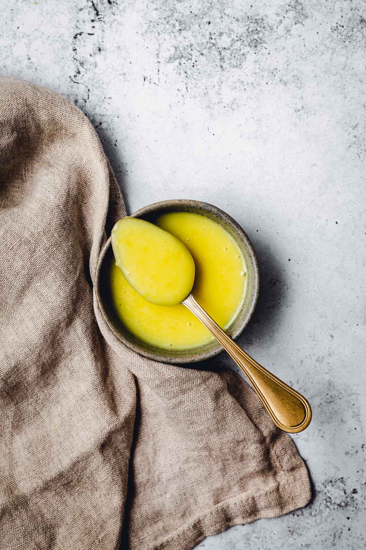 A small bowl containing lemon curd and a golden spoon with a piece of linen on the left side. 