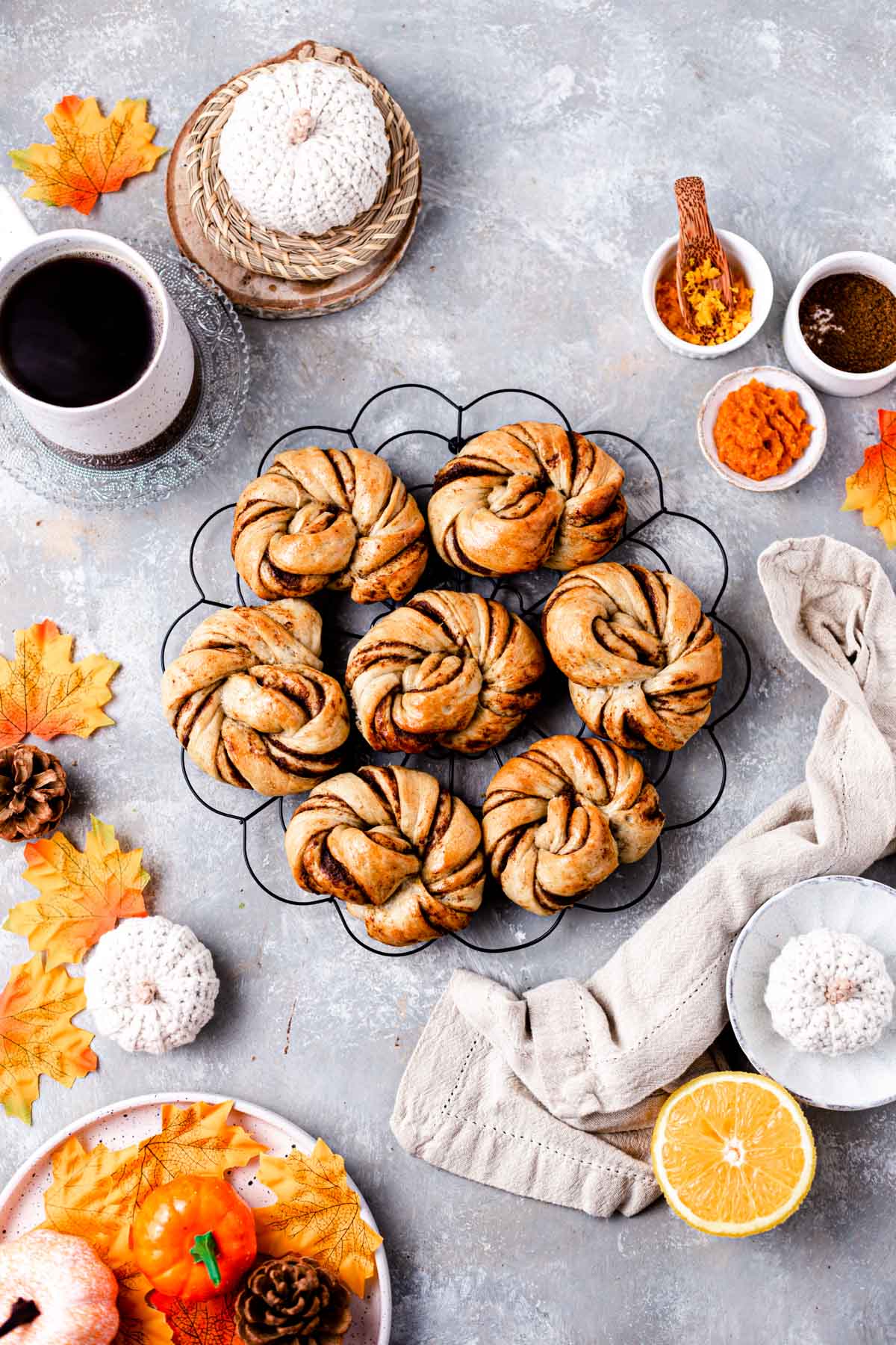 Seven pumpkin rolls placed on a round cooling rack and surrounded by orange leaves, coffee cup, pumpkin puree and more. 