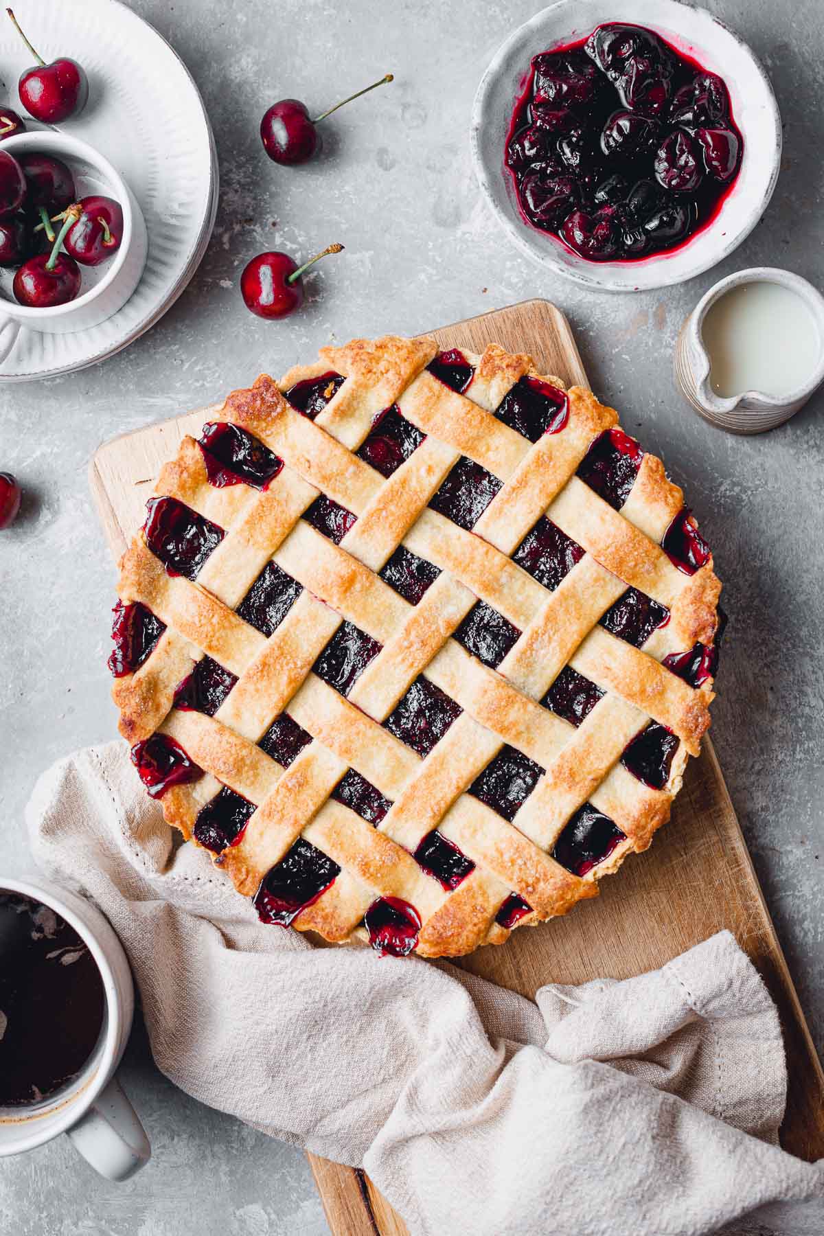 A baked vegan cherry pie placed on a wooden board surrounded by cherries and coffee. 