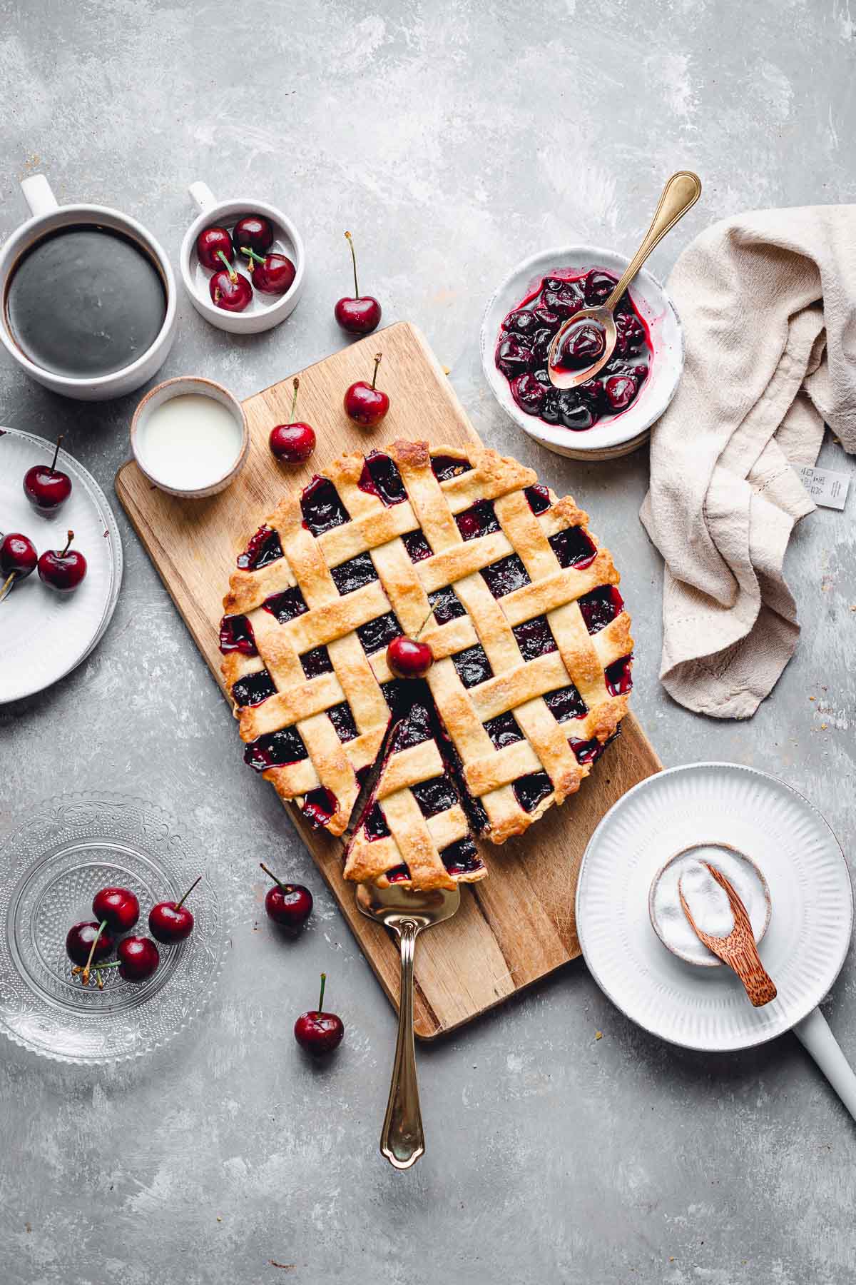 A cherry pie placed on a wooden board with various plates and bowls placed next to it.