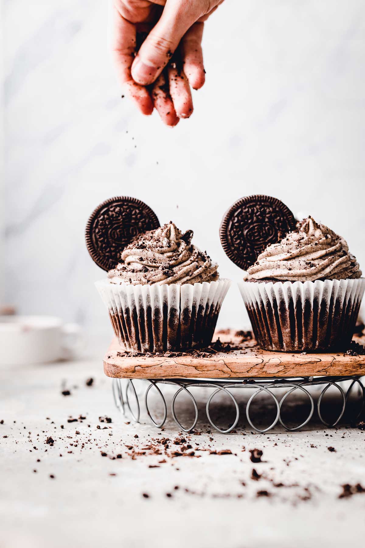 Two Oreo cupcakes placed on a cooling rack with a hand sprinkling additional Oreo powder on top.