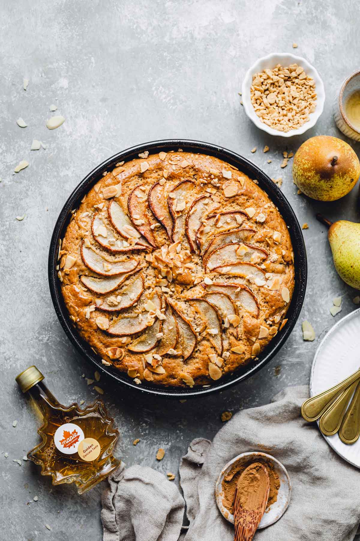 A round cake in a black tin surrounded by plates, bowls and pears. 