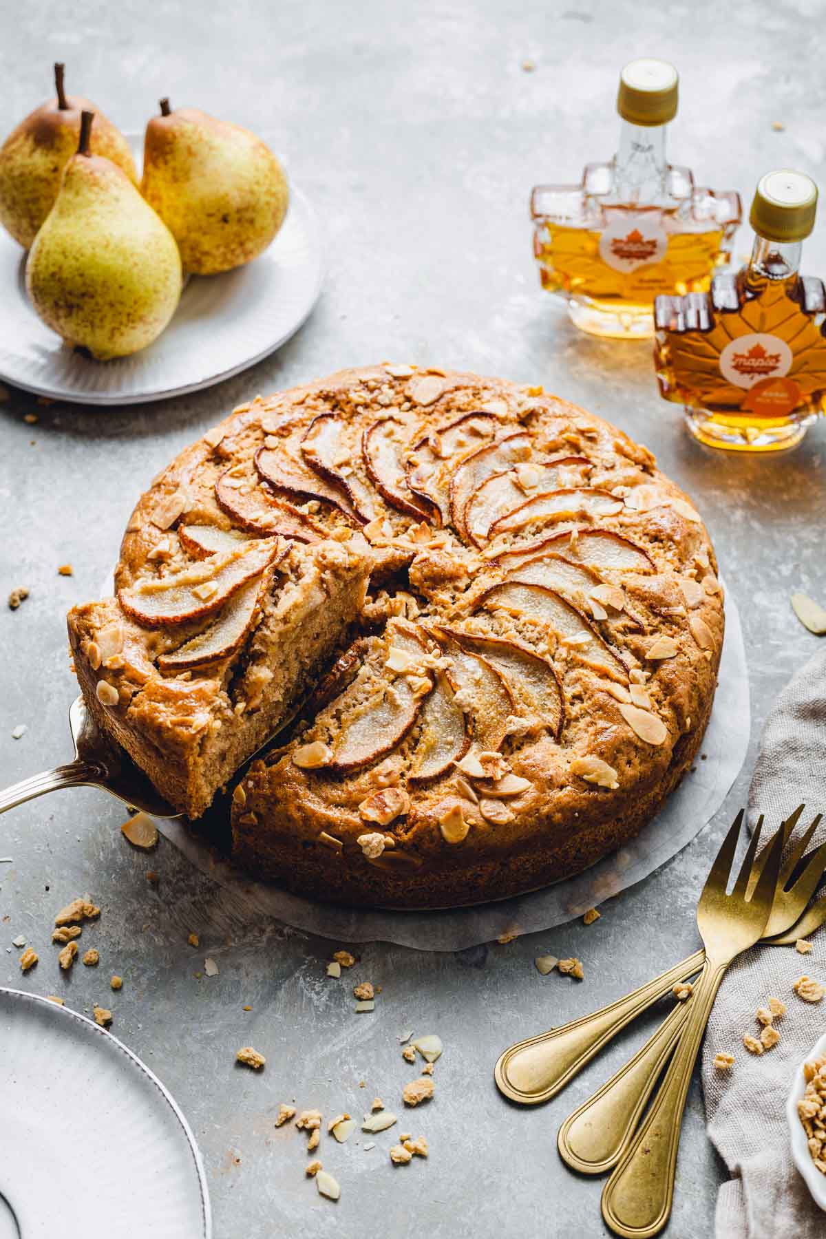 A side view of a round pear cake with various props like cutlery and plates next to it and a slice being taken out. 