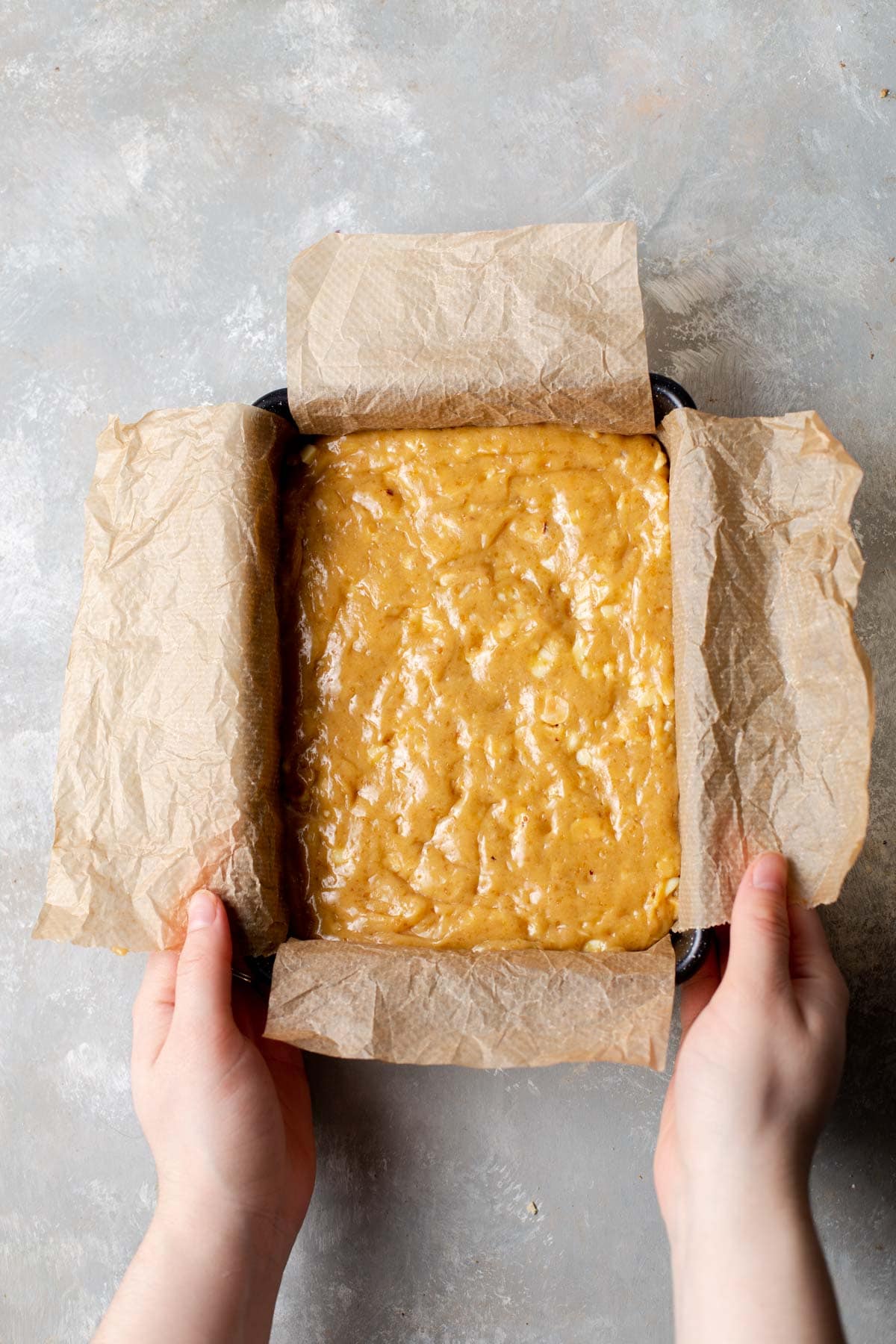 Two hands holding the sides of a lined baking tray with batter inside. 