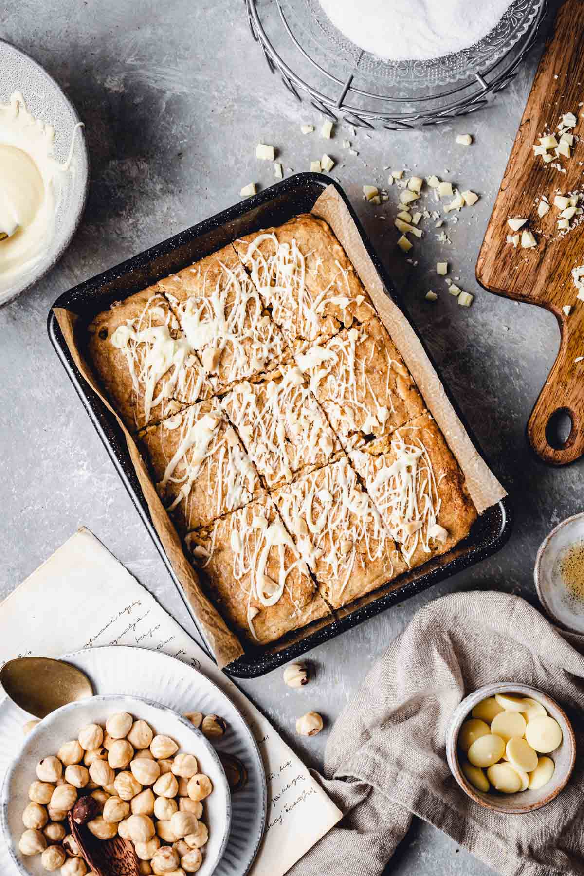 An overhead view of a blondie in a baking tin surrounded by various plates, bowls and boards. 