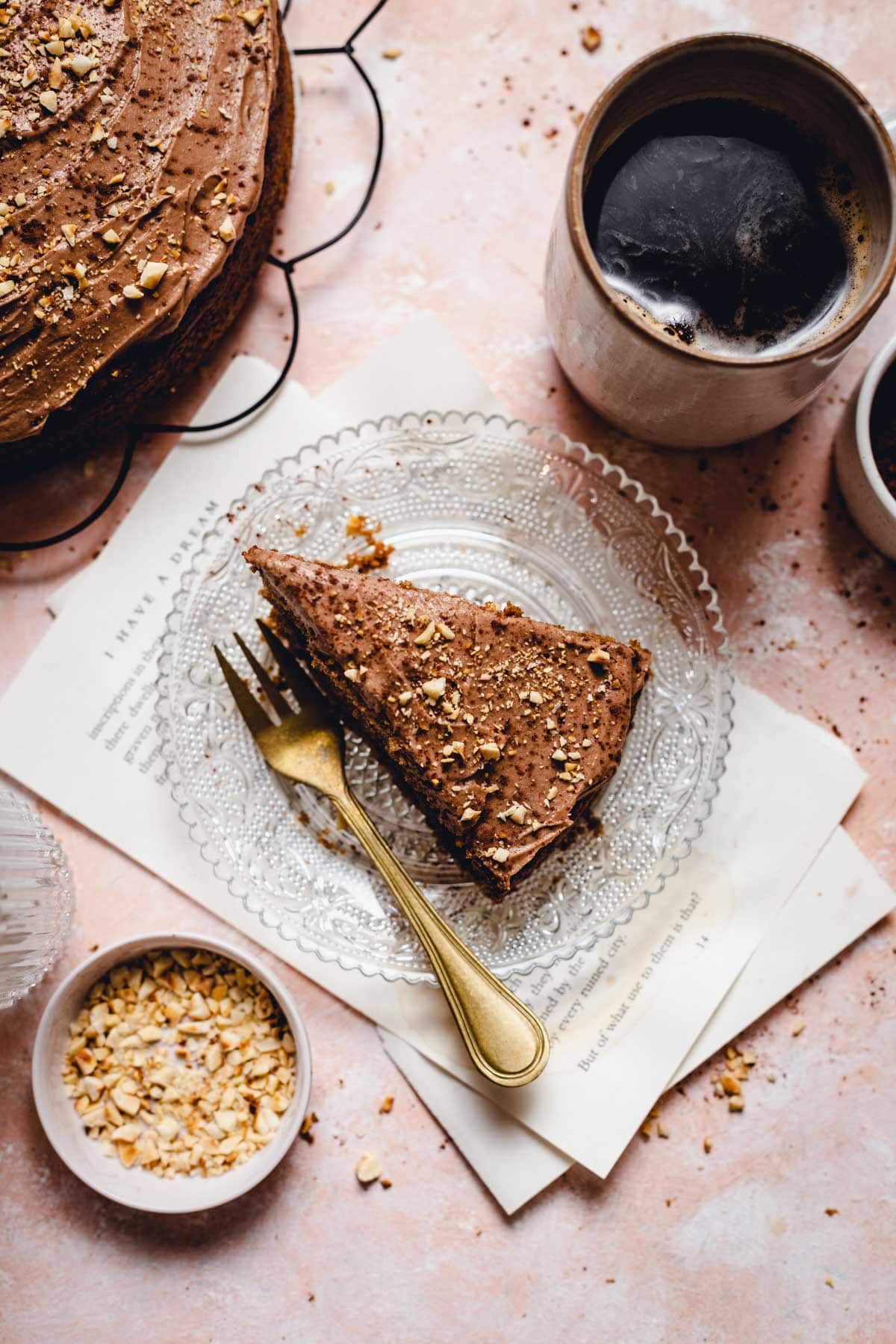 A slice of cake on a round glass plate with a gold fork and a cup of coffee next to it. 