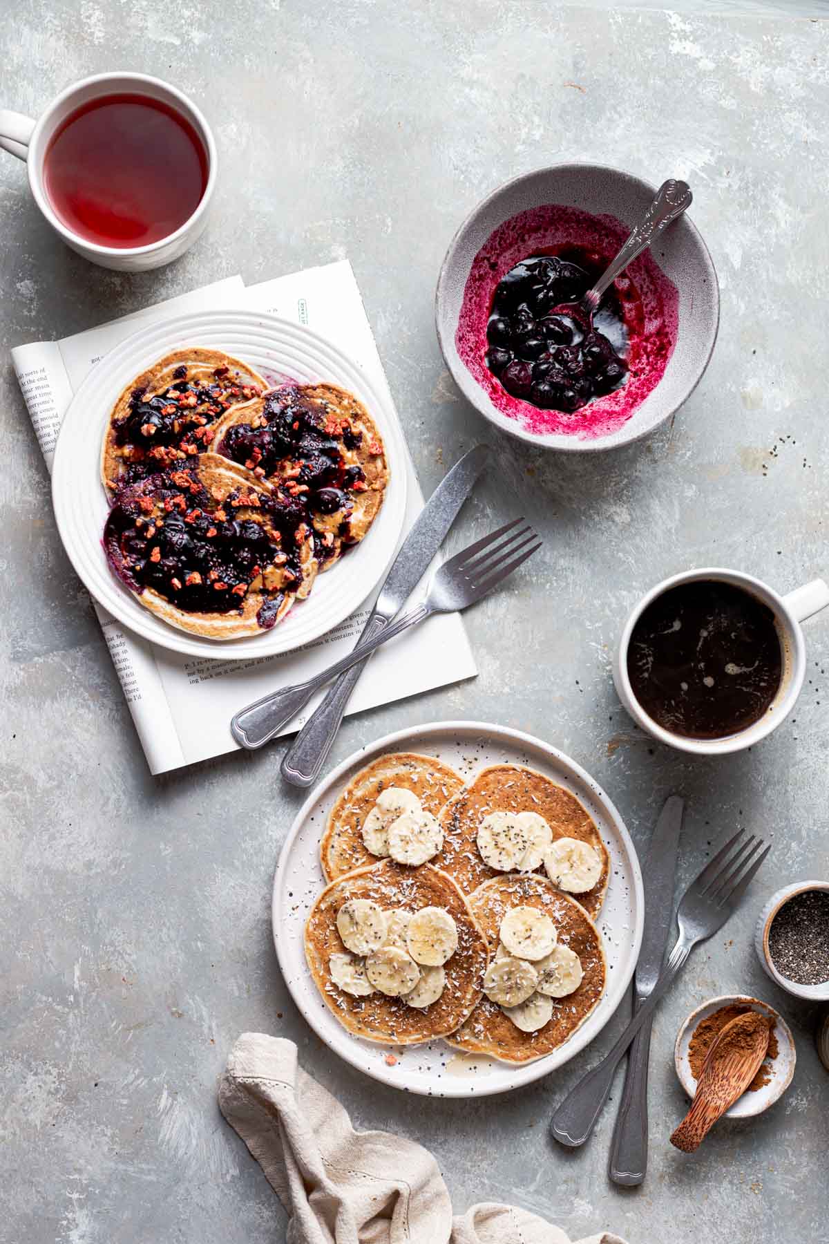 Two white plates of pancakes placed diagonally next to each other with mugs and bowls placed around them.