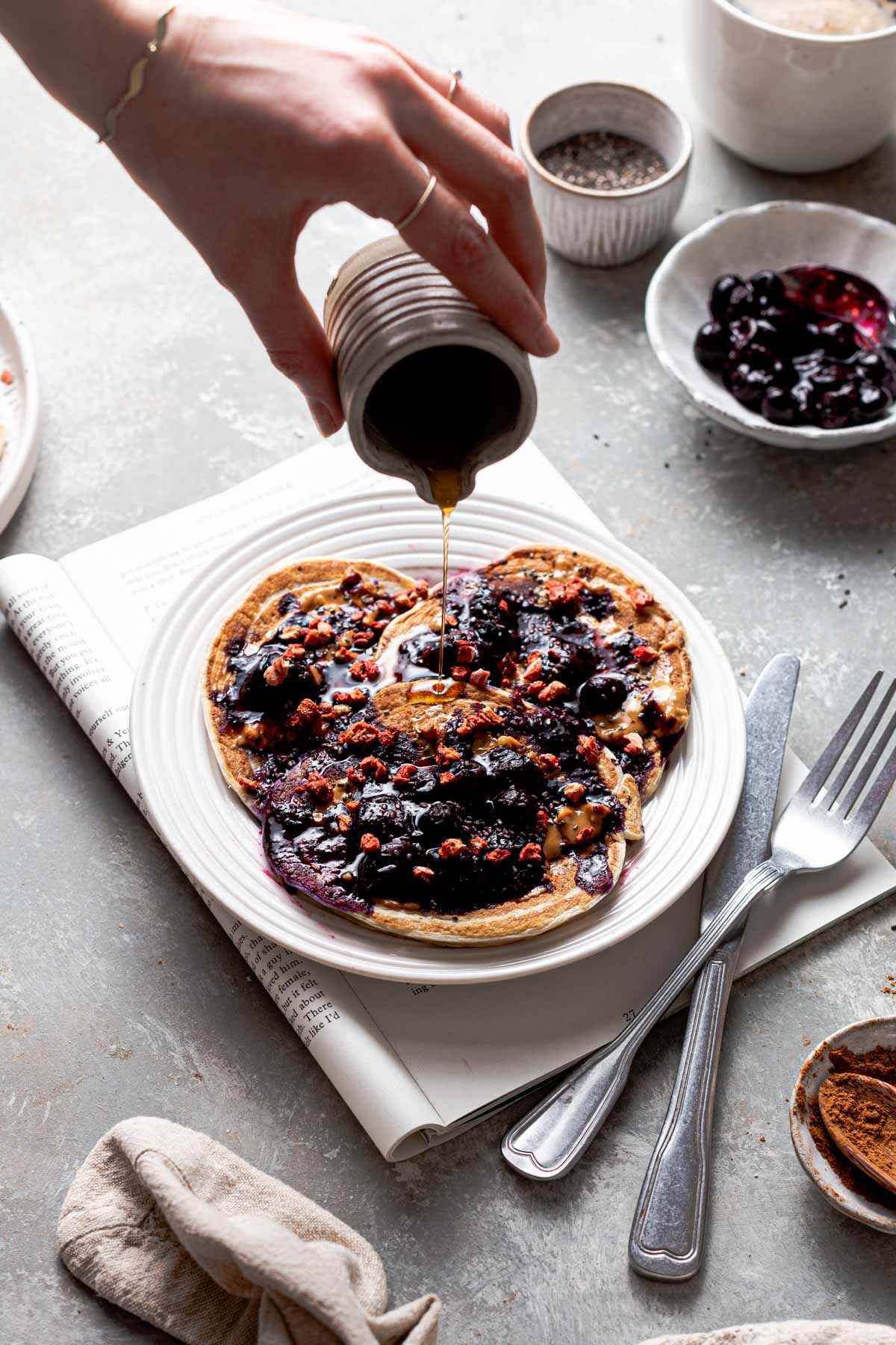 A hand pouring maple syrup over a plate of blueberry pancakes. 