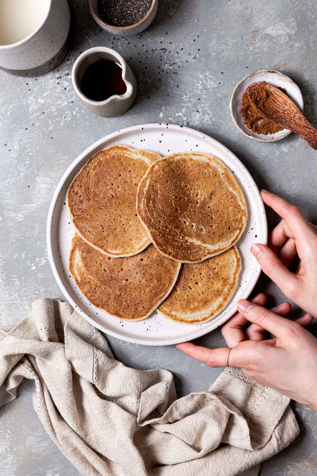 Four plain vegan pancakes on a white plate with two hands holding the sides and a piece of linen next to it. 