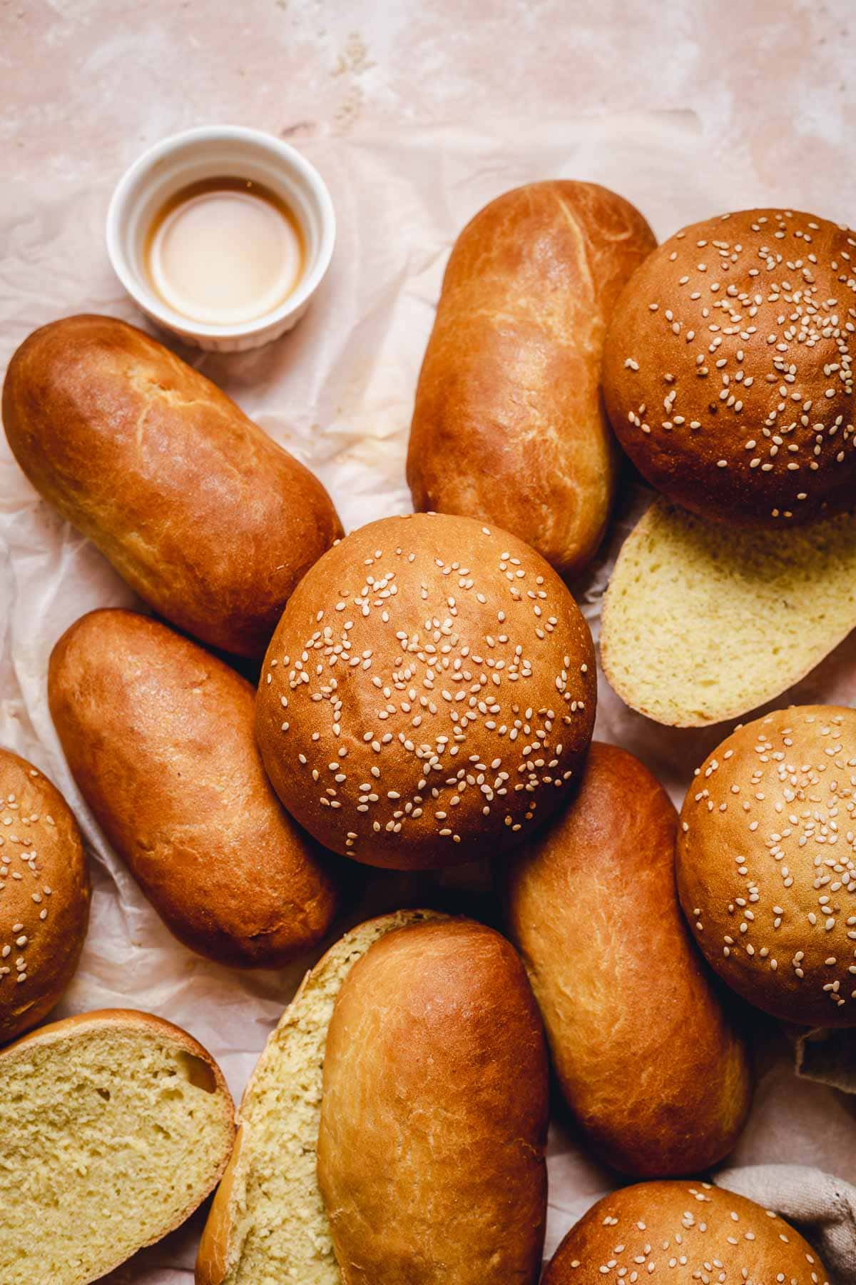 Several bread buns and hotdog buns brushed with vegan egg wash and placed on top and next to each other on crumpled baking paper.