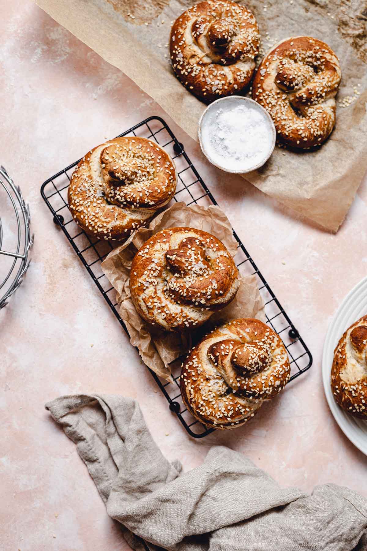 Sesame pretzels on a peach coloured surface with racks, napkins and baking paper in sight.