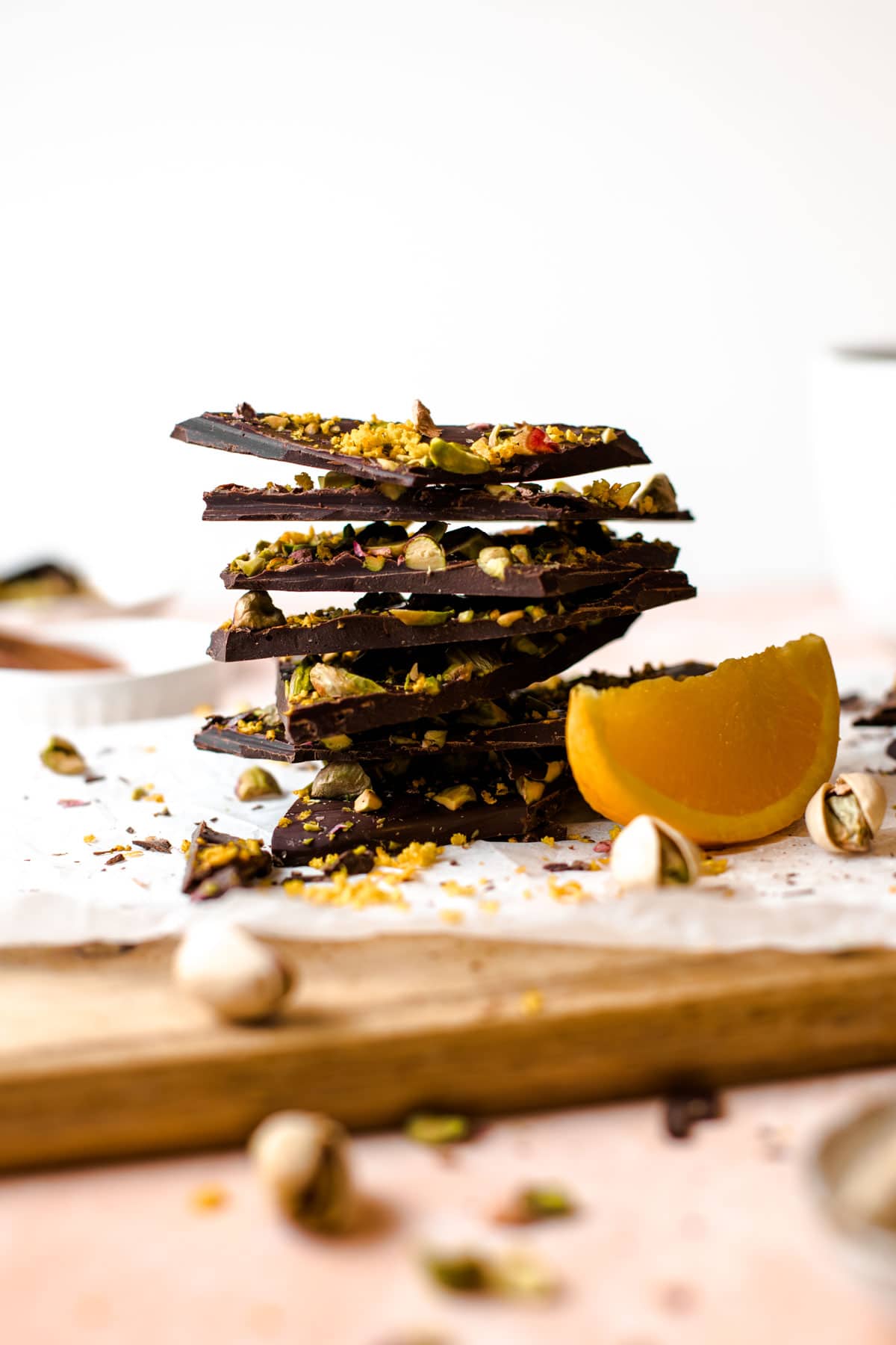 A side view of pieces of chocolate bark placed on a wooden chopping board and a slice of orange placed next to it. 