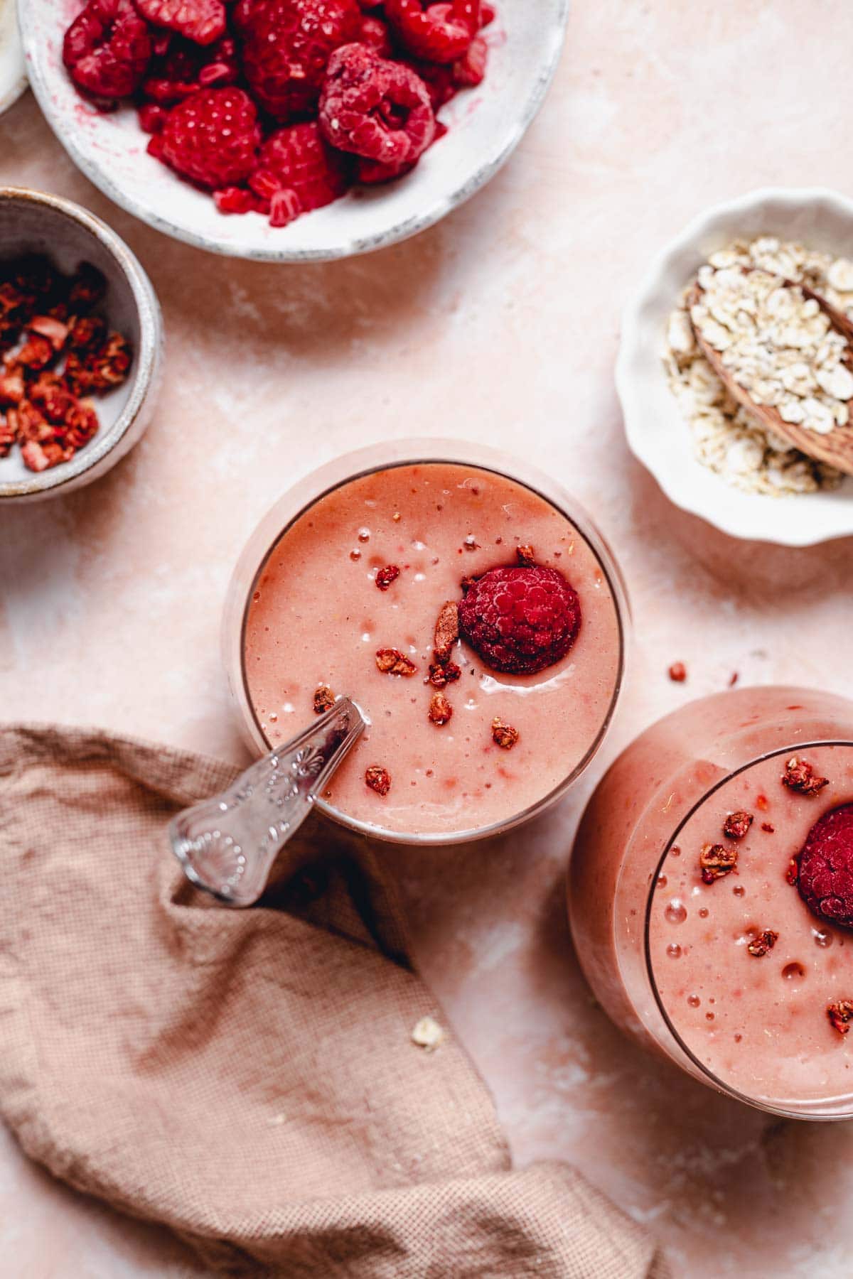 Two glasses of pink coloured smoothie on a flat background with raspberries, oats and a napkin laid next to them.