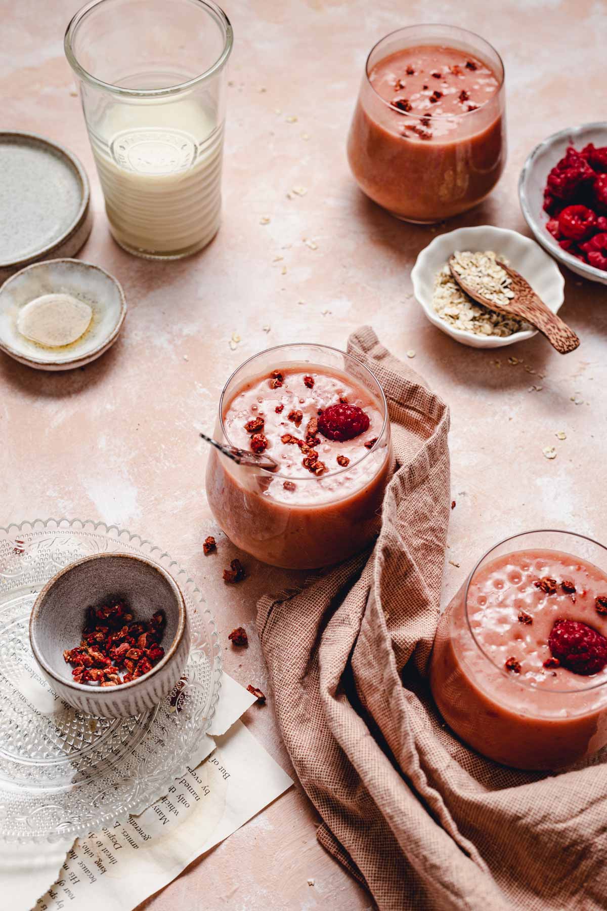 A side view of three glasses of oat milk smoothie placed on a pink surface with a piece of linen and various props around it. 