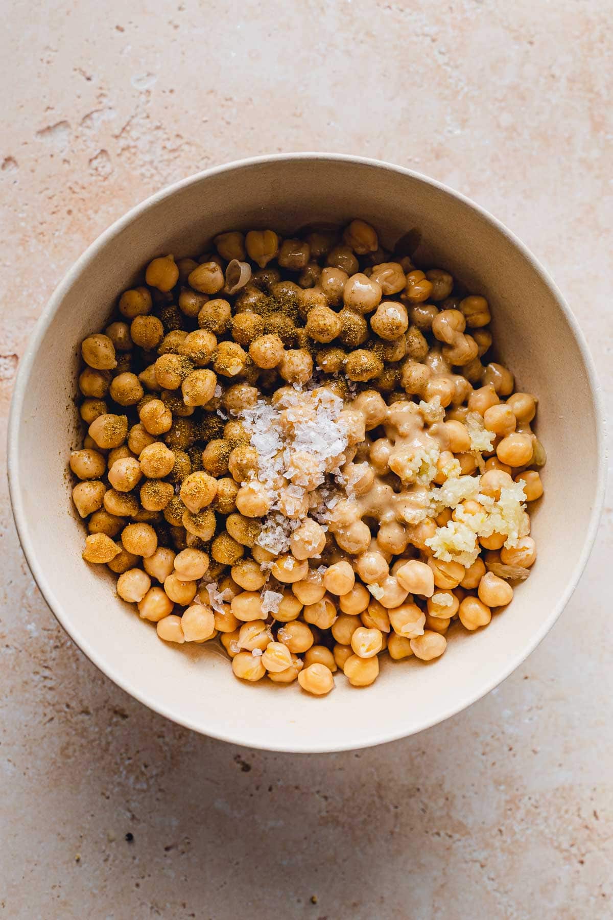 A large bowl with chickpeas, salt, minced garlic and other hummus ingredients prior to blending