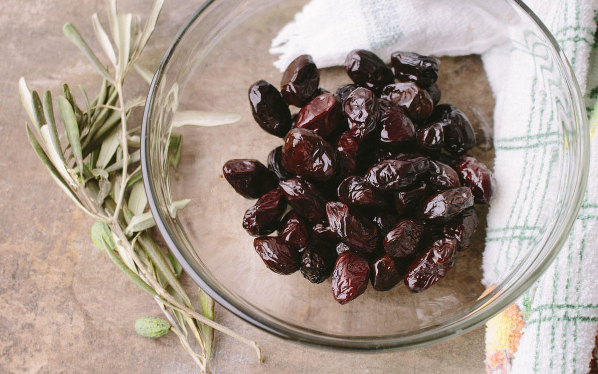 Black olives in a large glass jar with some olive leaves and a piece of linen around it.