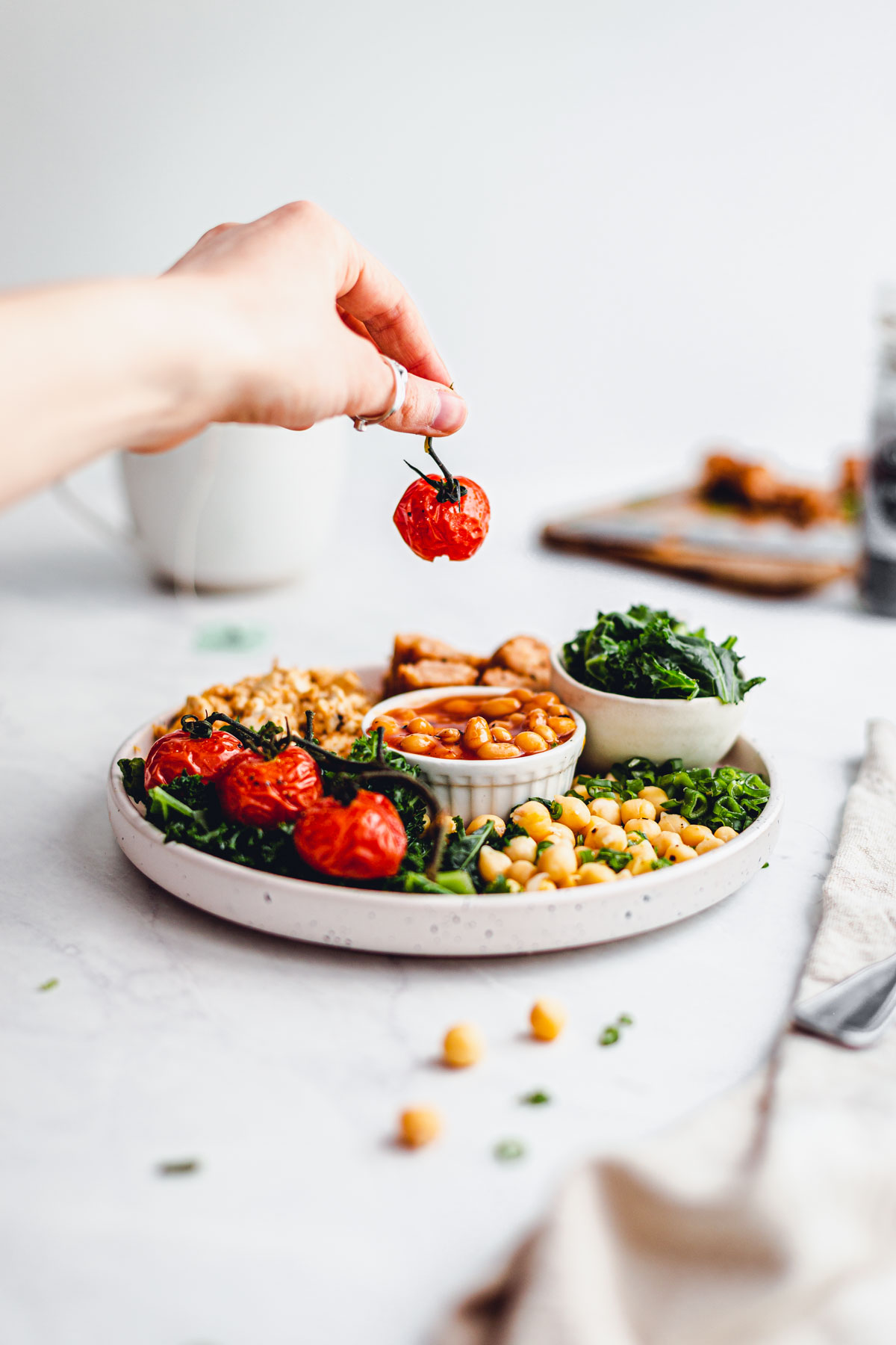 A hand reaching out to place a cherry tomato on a plate filled with vegan English breakfast.