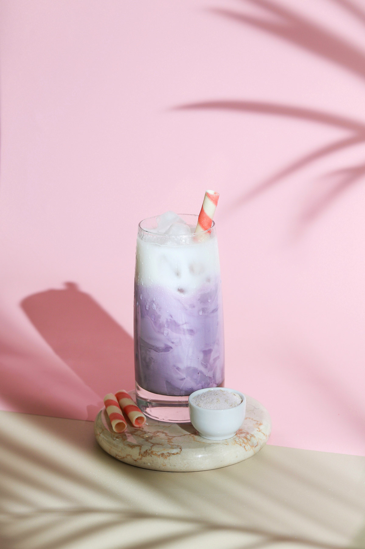 A glass of taro milk tea with ice cubes placed on a marble coaster in front of a pink background.