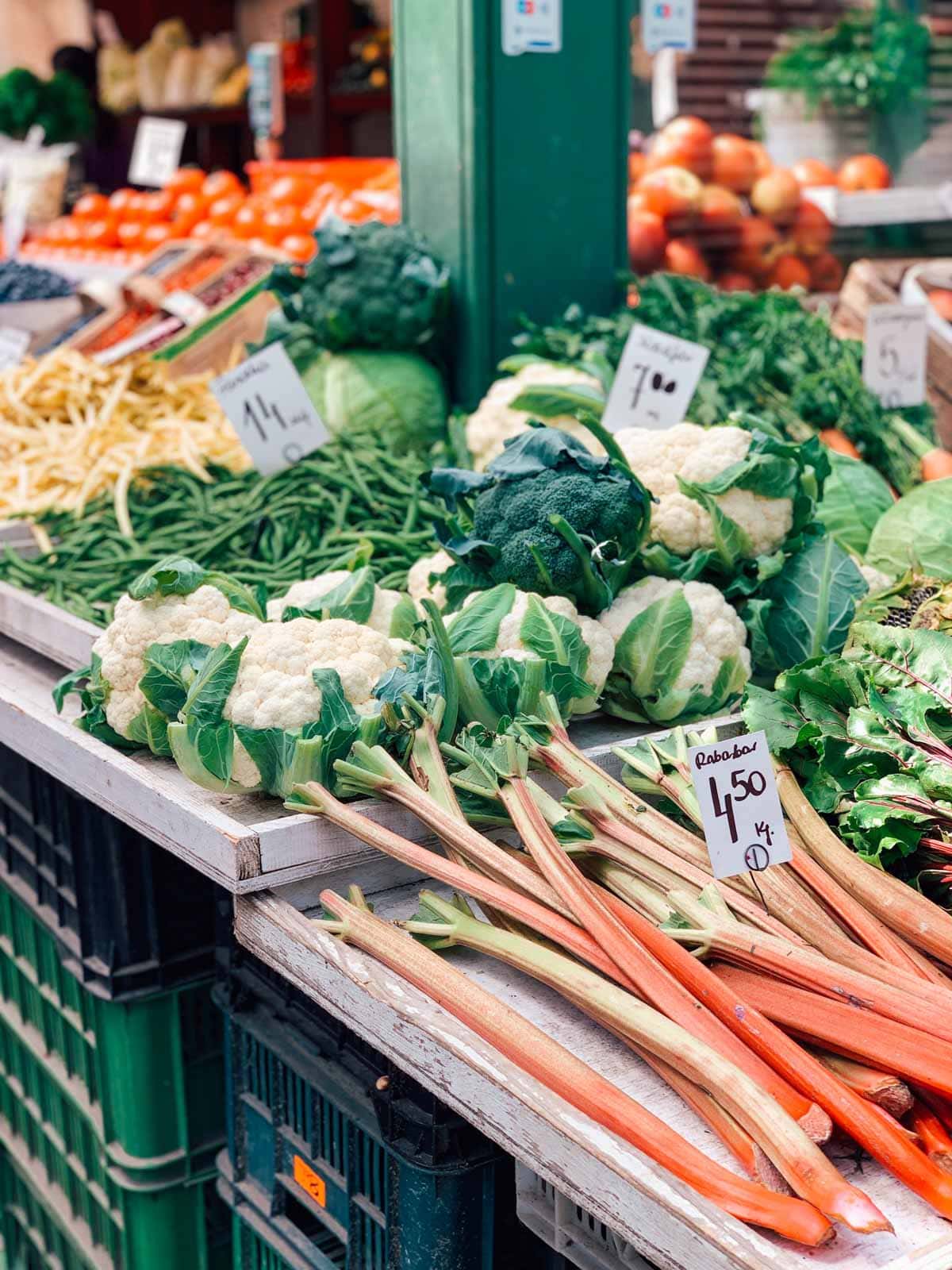 A large market stall with various vegetables including rhubarb, cauliflower, and beans.