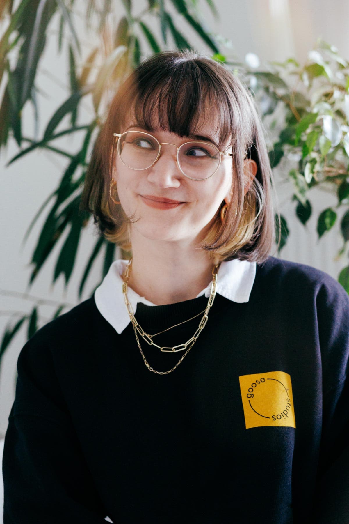 A portrait of a woman smiling and looking to the side wearing a black jumper and featuring many plants in the background. 