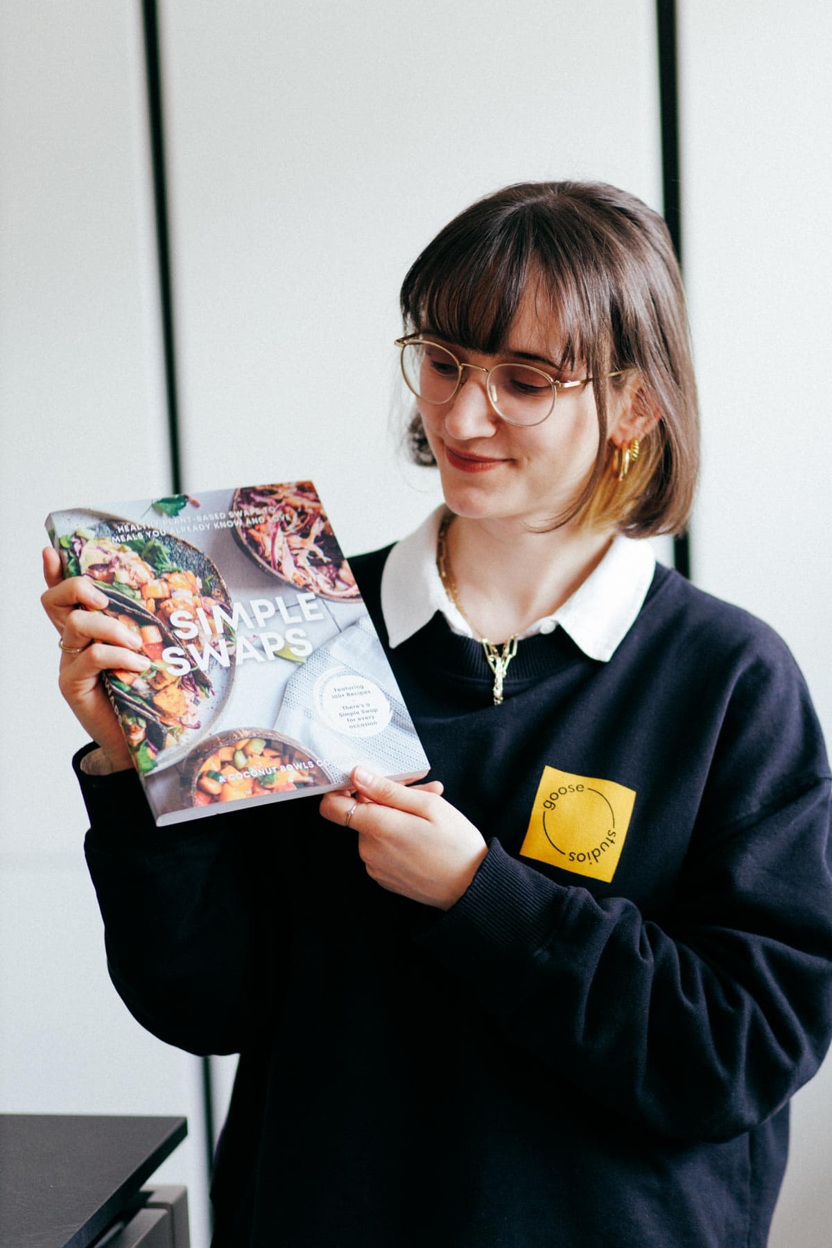 A woman holding out a cookbook and looking at it while wearing metal glasses and a black jumper.