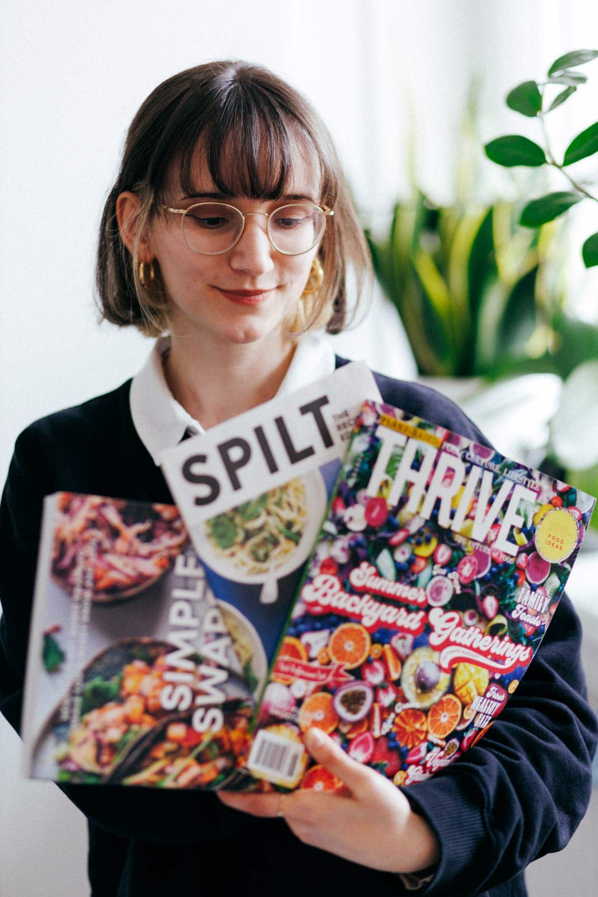 A woman holding three magazines and looking at them with plants in the distance. 