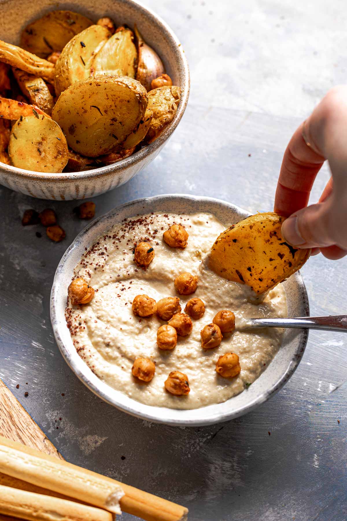 A hand reaching out to dip a spicy potato wedge into a bowl of hummus.