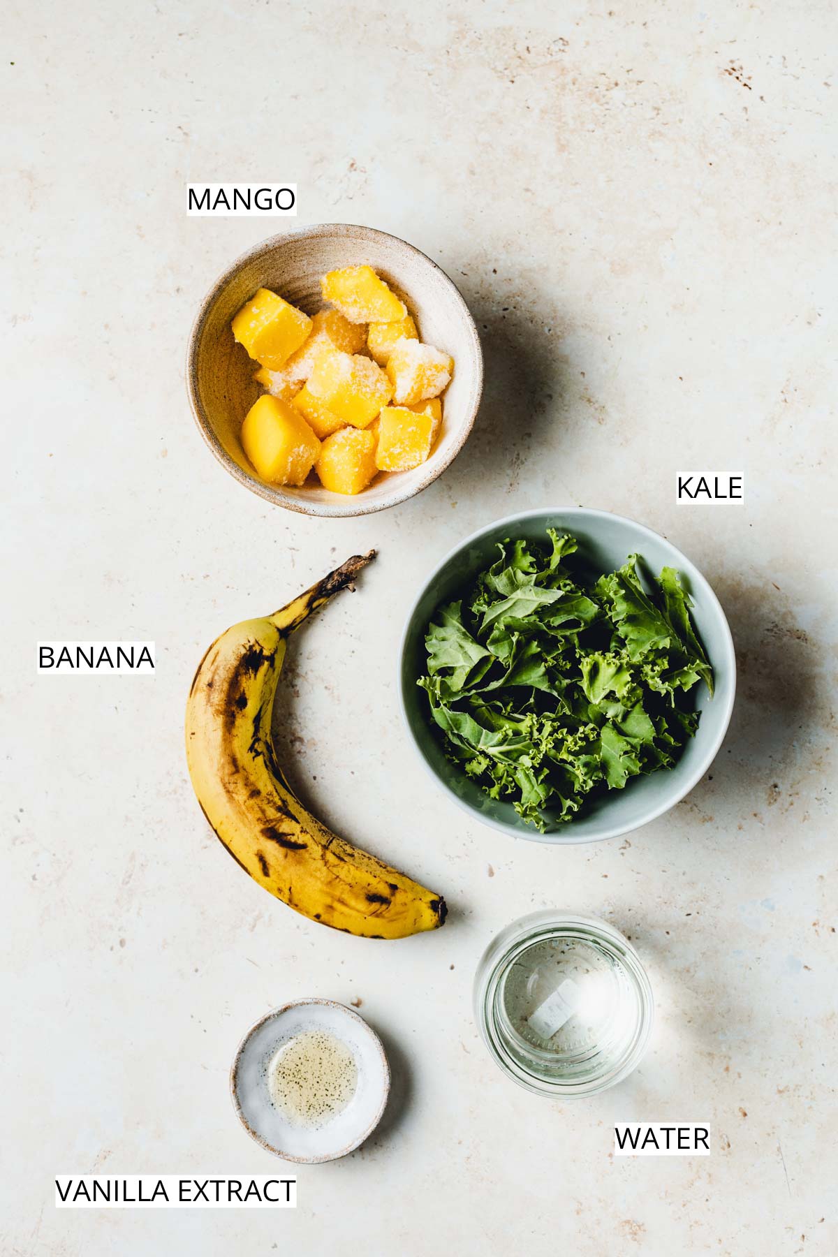 Bowls of mango, kale, vanilla extract, water and banana placed next to each other on a flat surface.