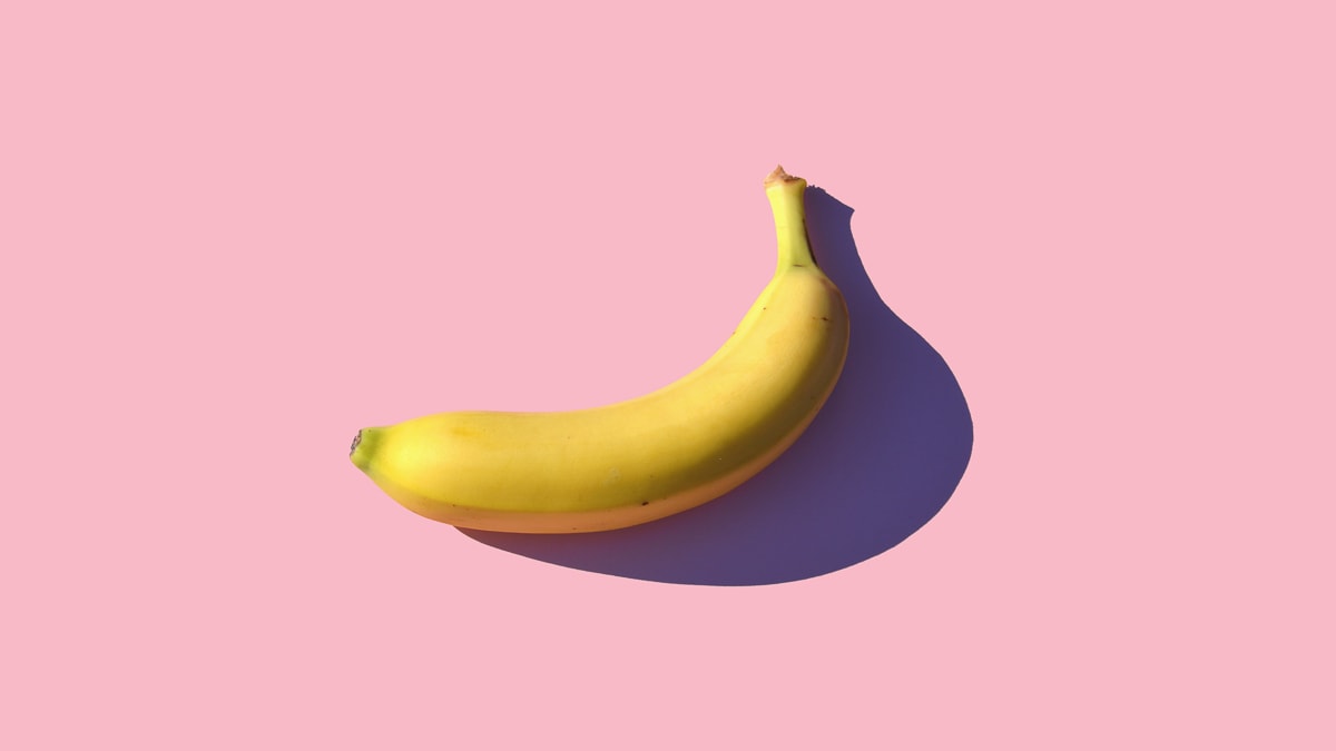 A single banana laid flat on a plain pink surface.