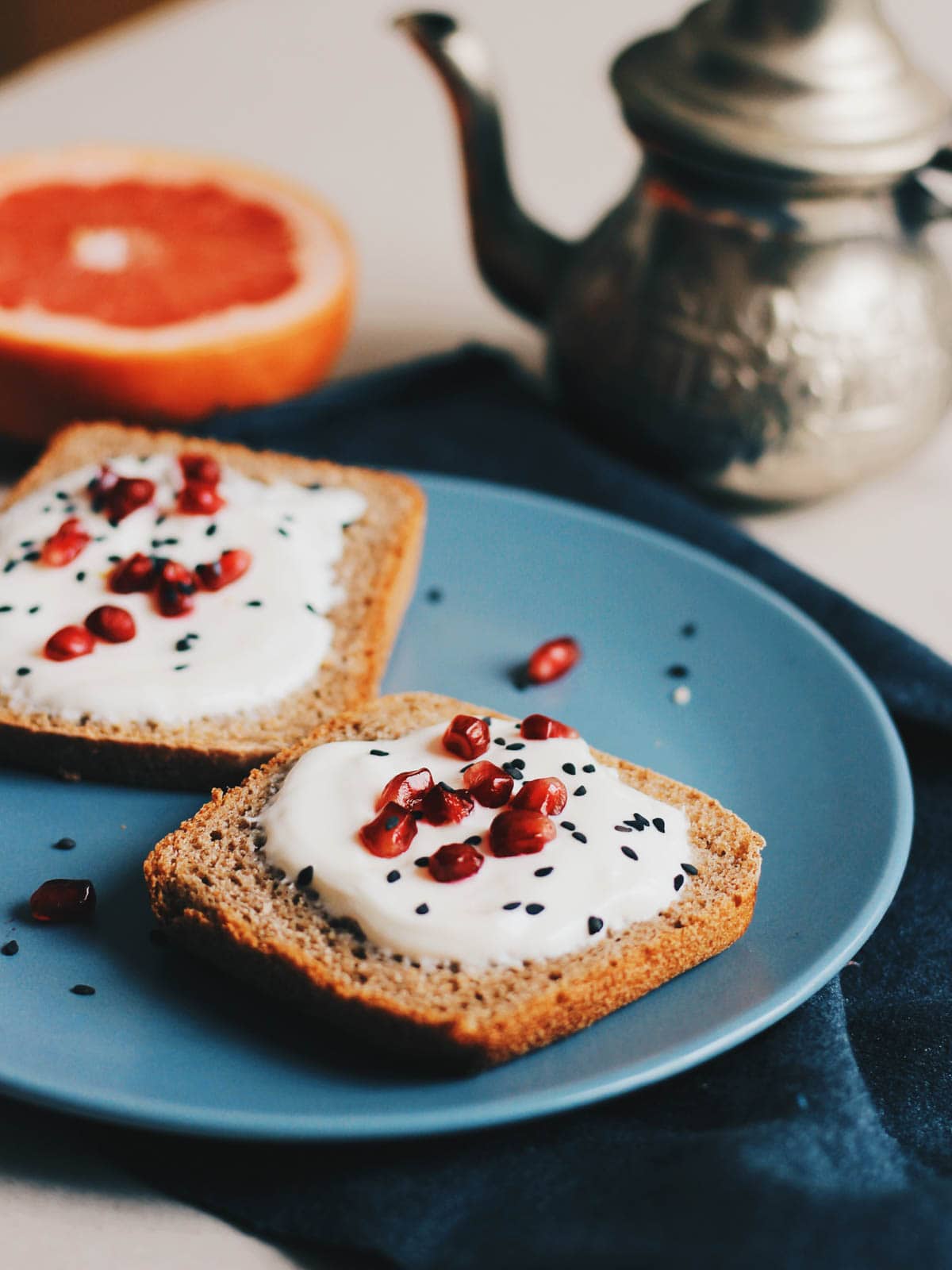 Two pieces of toast placed on a blue plate with a creamy topping, pomegranates and nigellas seeds sprinkled on top.