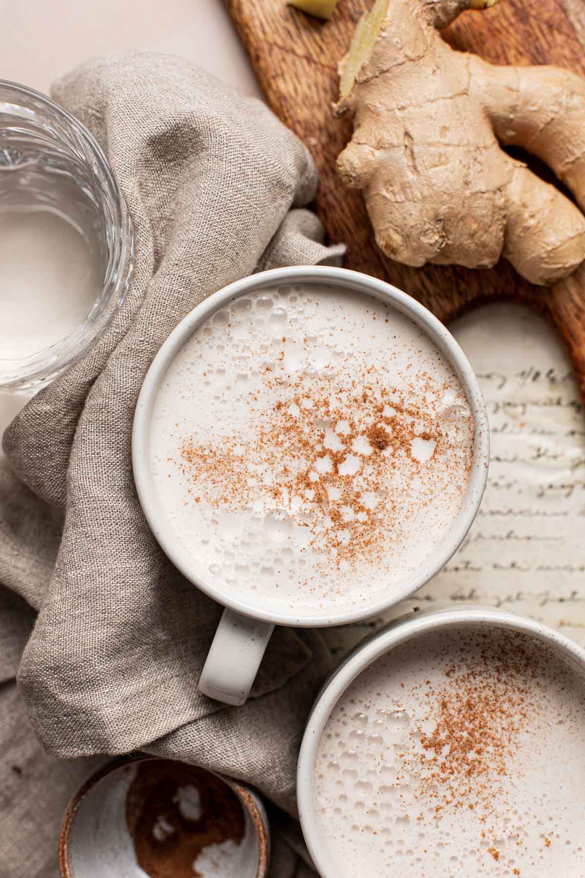 Two mugs containing a frothy milk drink with a ginger root placed next to them.