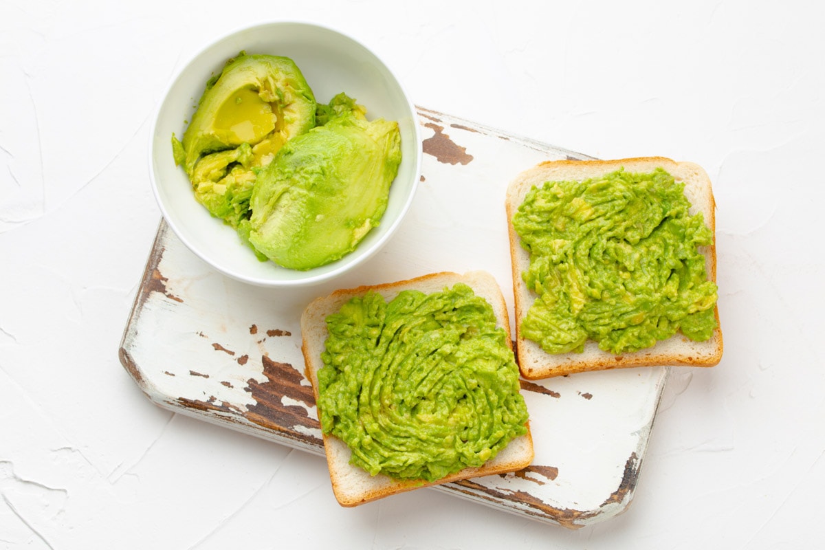 Avocado toasts and smashed fresh ripe avocados in bowl on wooden rustic cutting board.