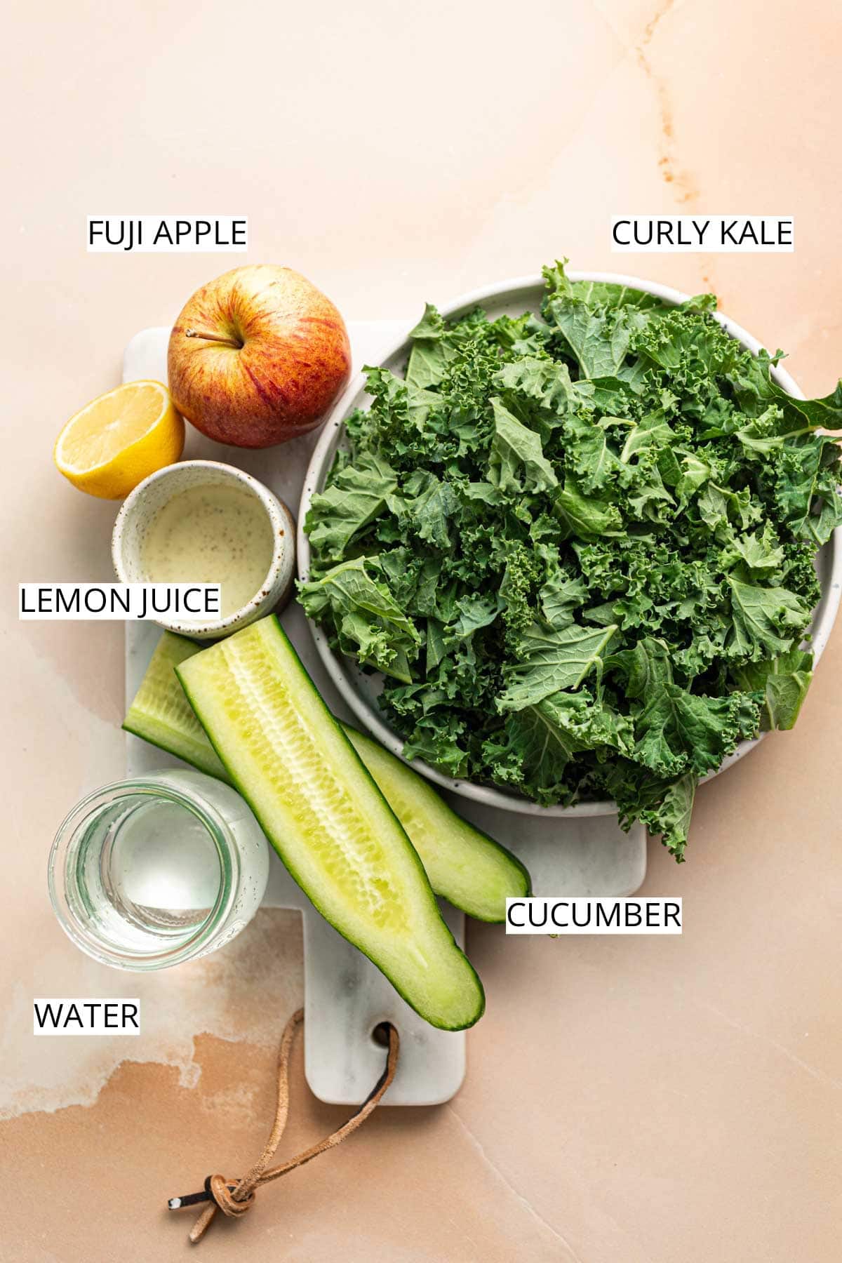 Kale, cucumber, lemon juice, water and apple on a marble cutting board.