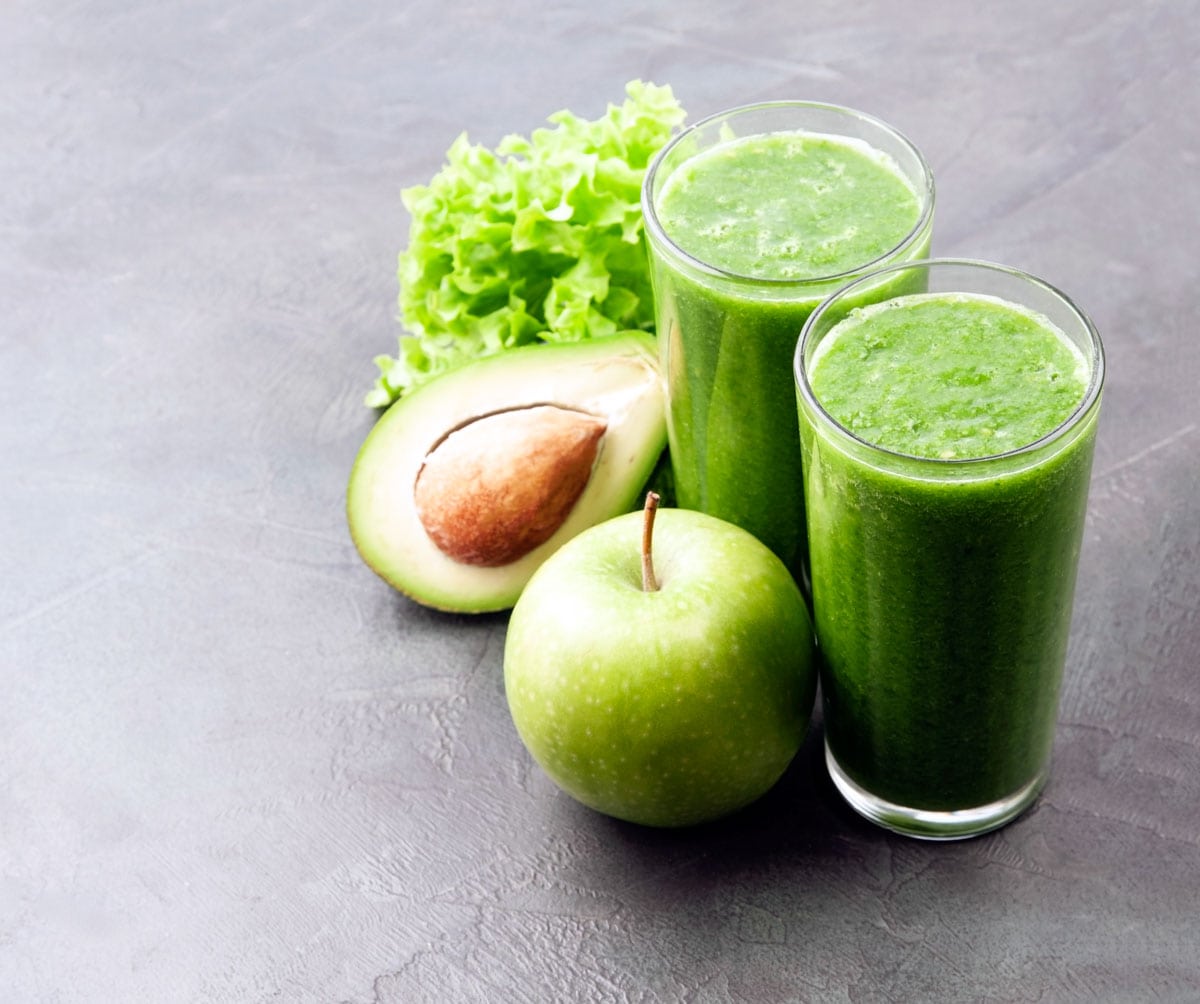 Two green smoothie glasses with lettuce, avocado and green apple placed next to them.